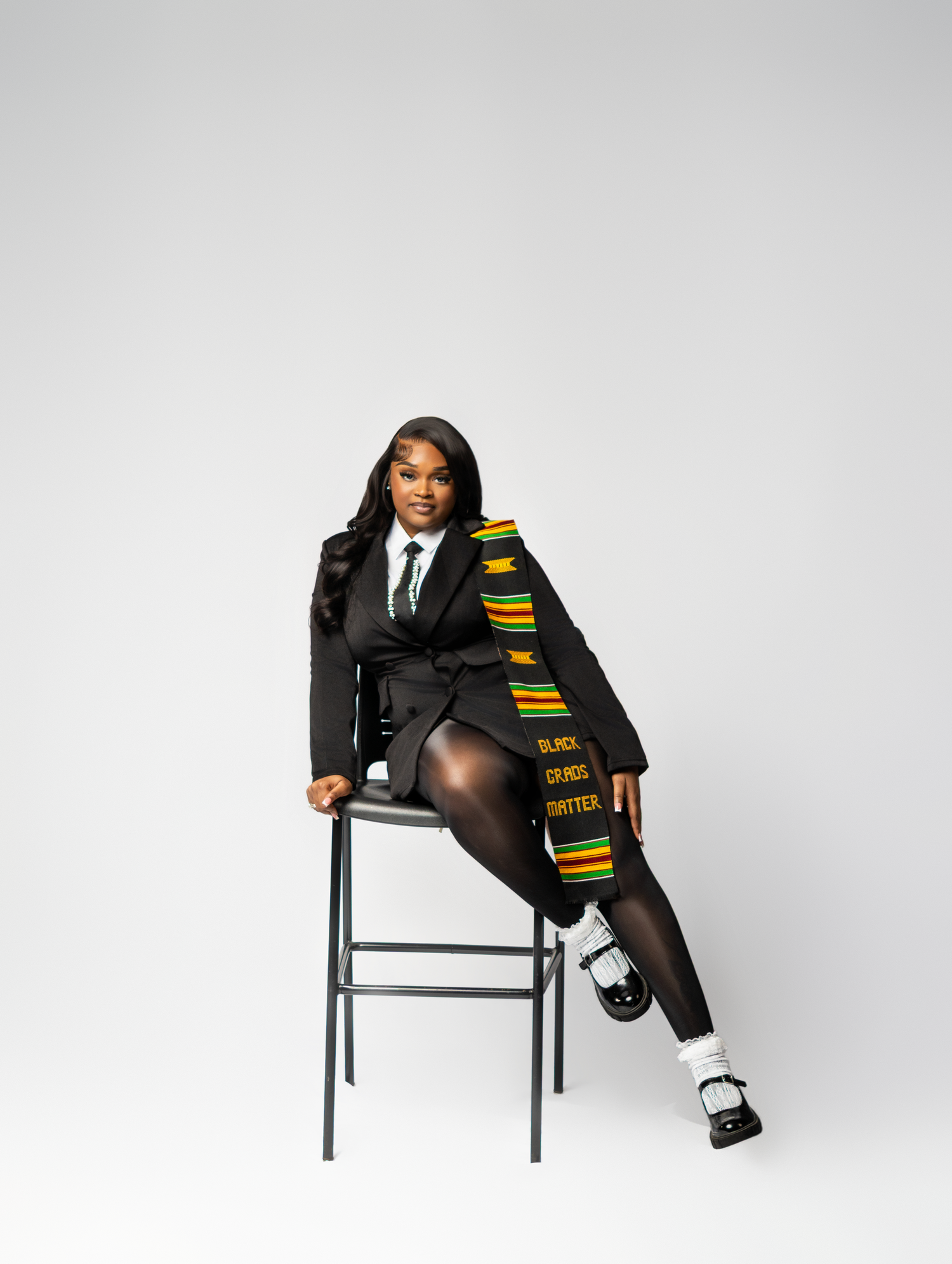 Graduate poses on stool wearing black suit with kente cloth Black Lives Matter stole and Mary Jane shoes.