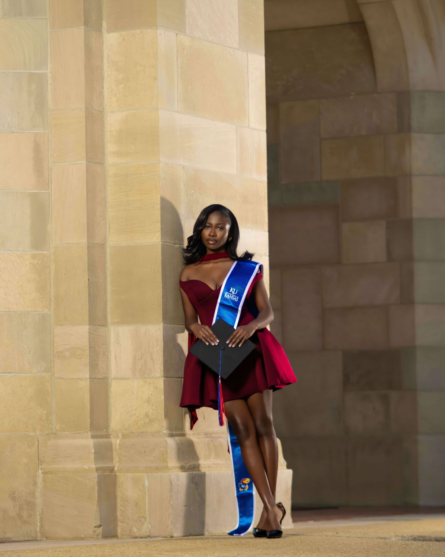A young woman in a red dress and blue graduation sash poses with a cap at a stone university building.