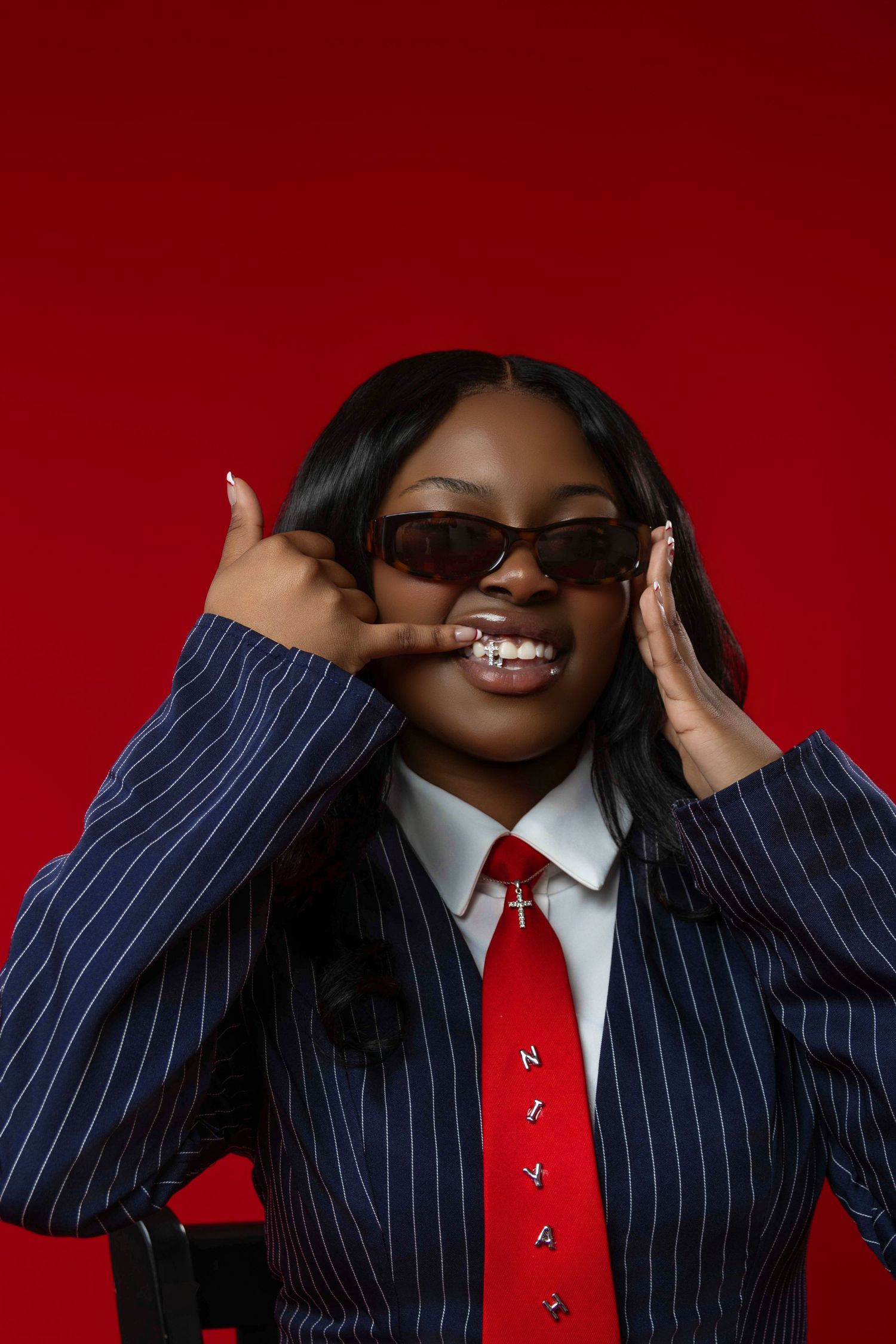 Young Black woman in pinstripe blazer and red tie poses with sunglasses against bold red background.