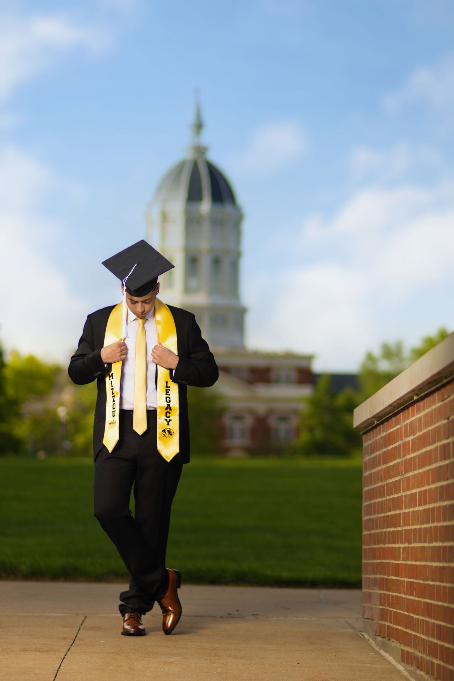 Graduate in black suit and gold stoles poses confidently near brick wall with domed university building in background.