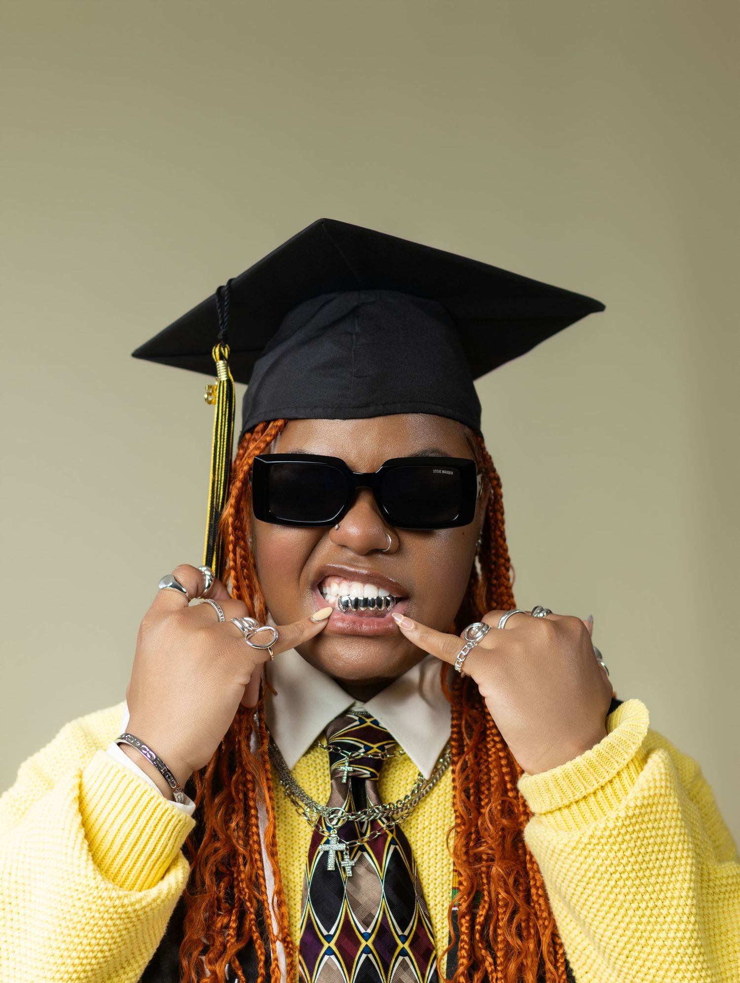 Graduate in yellow sweater, graduation cap, sunglasses, and grillz posing with fingers on teeth.