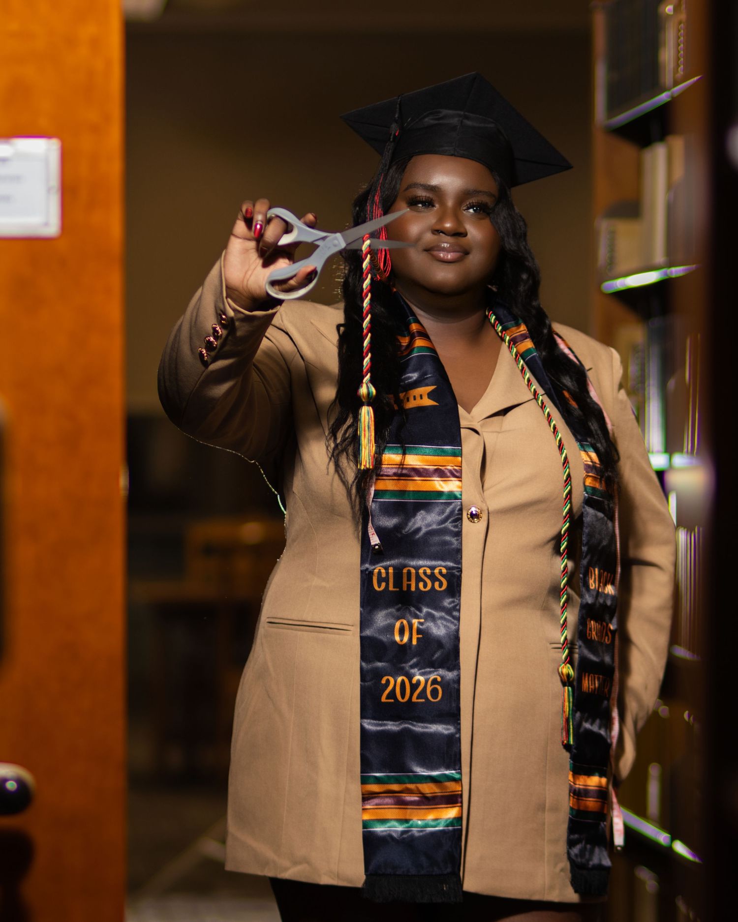 A smiling graduate in cap and Kente cloth stole reading Class of 2026 holds scissors toward camera in warm indoor setting.