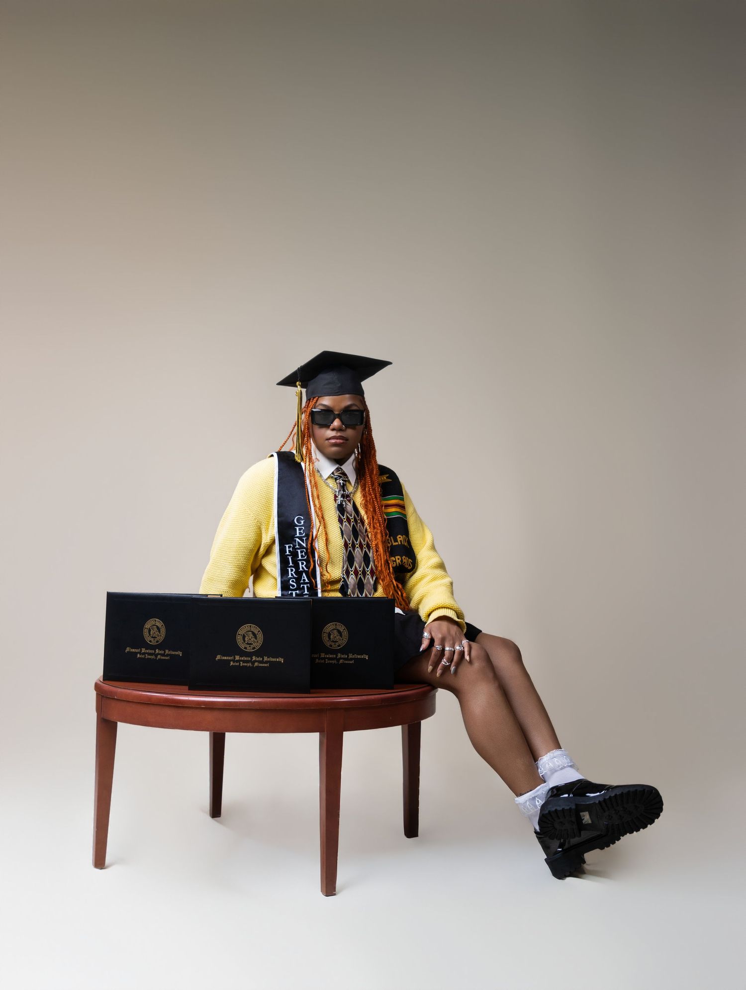 Graduate in yellow cardigan and cap poses with diploma boxes on a wooden table in a studio portrait.