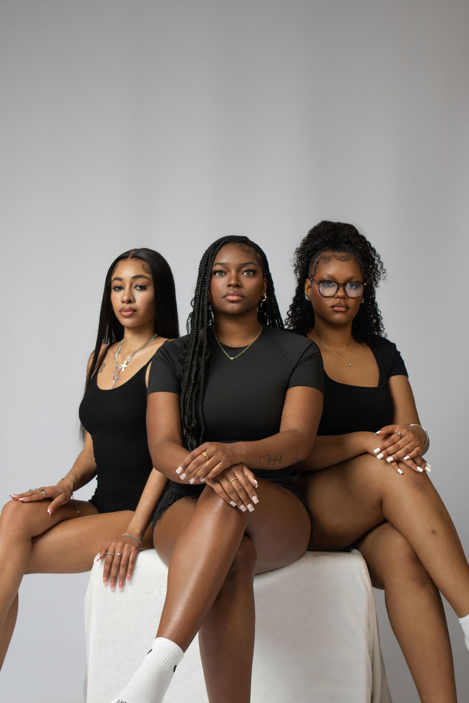 Three young Black women in matching black bodysuits pose confidently together against a gray studio background.