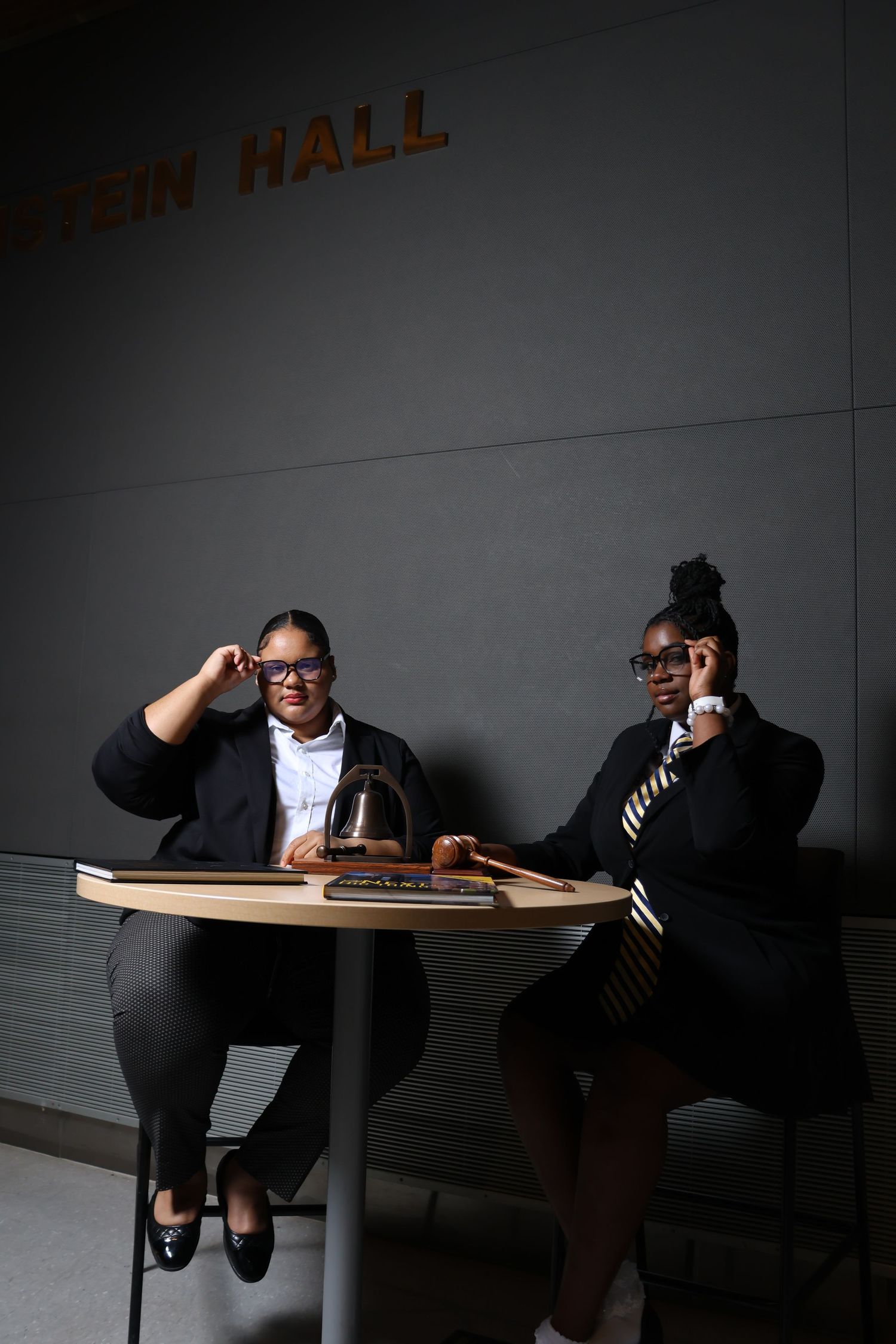 Two professional women in black suits sit at a table inside Stein Hall, one adjusting glasses.