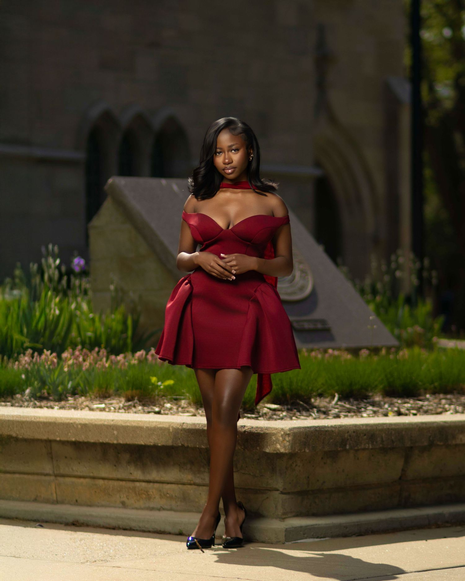 Woman in elegant red off-shoulder mini dress posing outdoors near stone monument in lush garden setting.