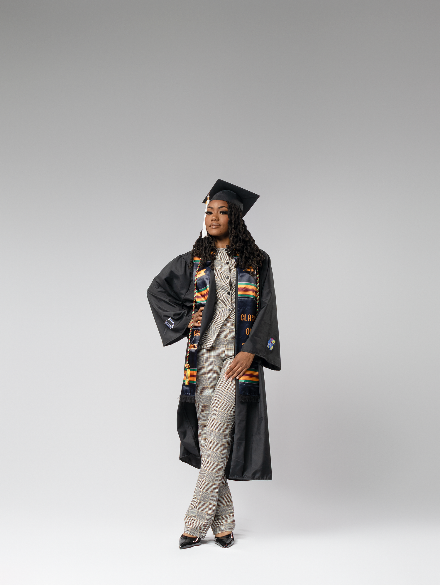 A young woman in a black graduation gown and cap with a colorful Kente stole poses confidently.