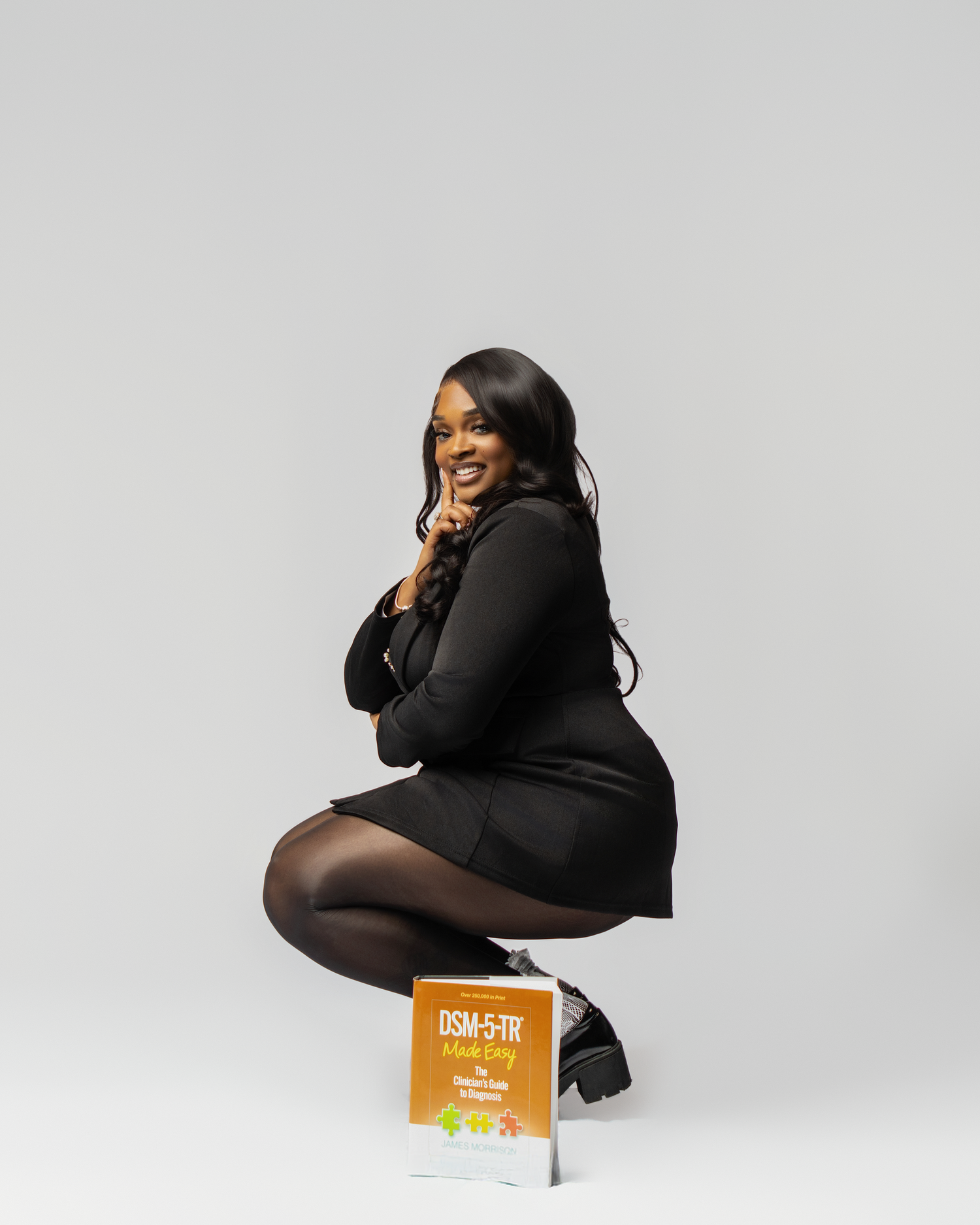 Woman in black dress squatting beside a DSM-5-TR book, smiling confidently against a gray background.