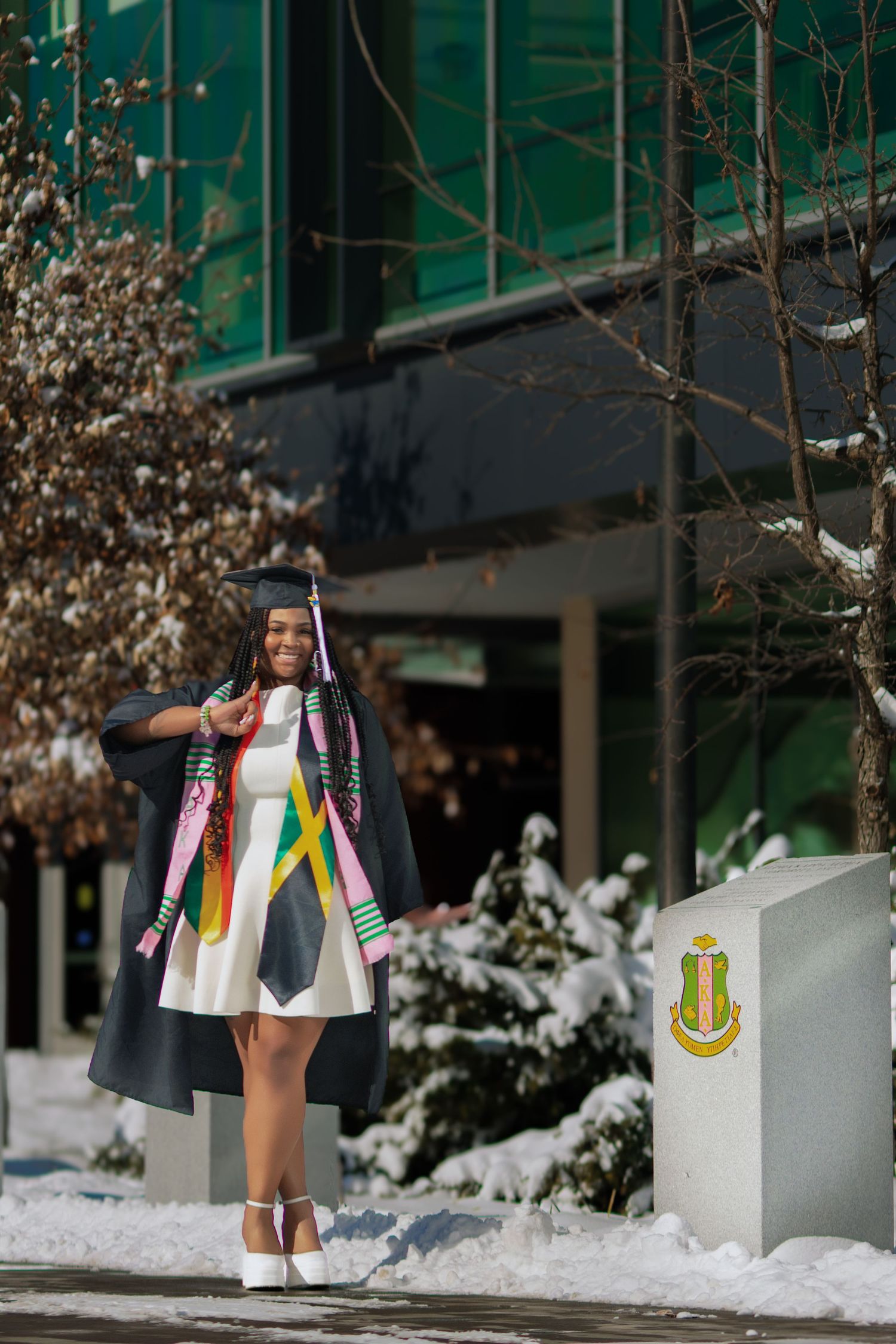 Graduate in cap and gown with kente stole poses proudly outside snowy university building.