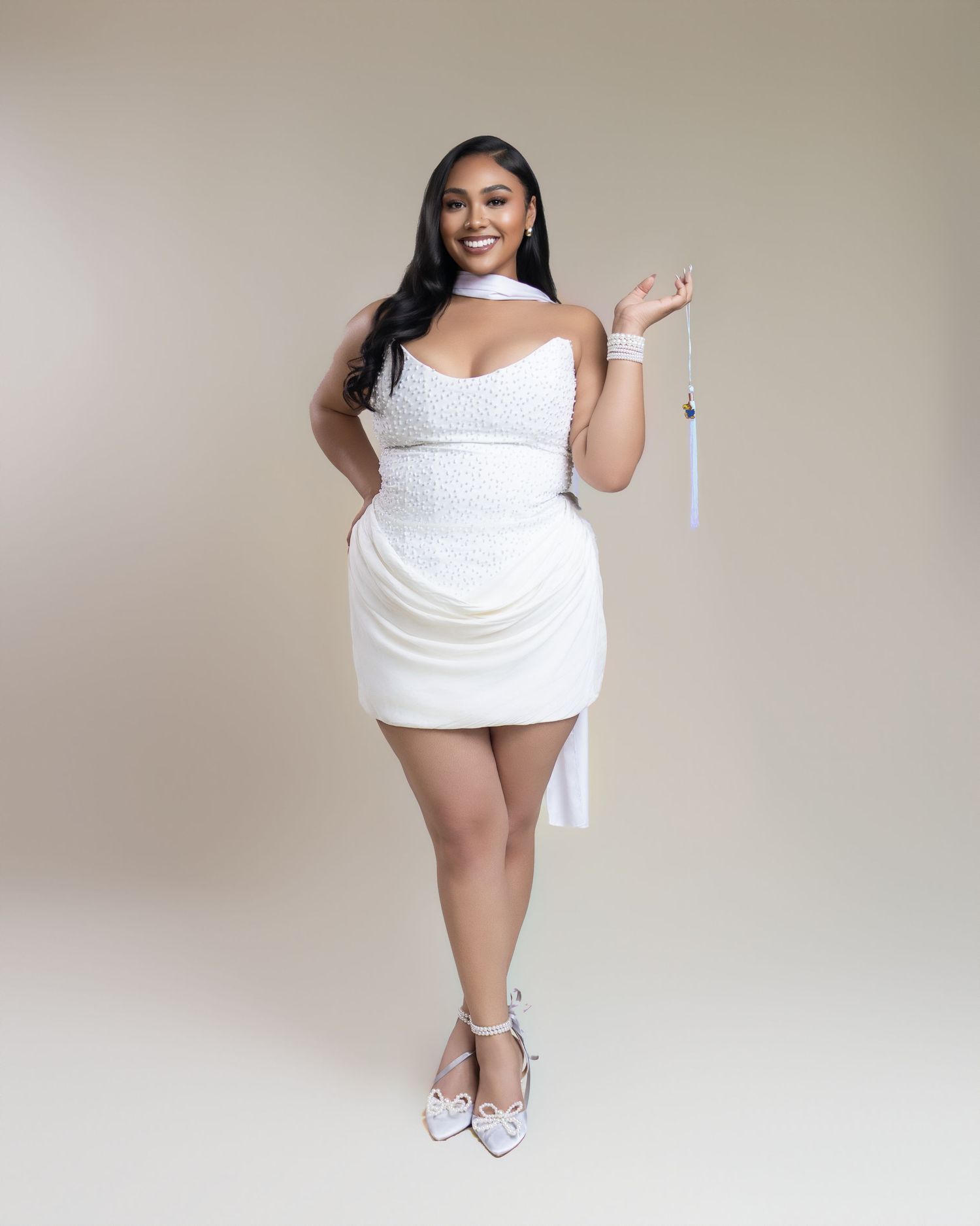 Woman in white mini dress and heels holding graduation tassel, posing confidently for graduation photos.