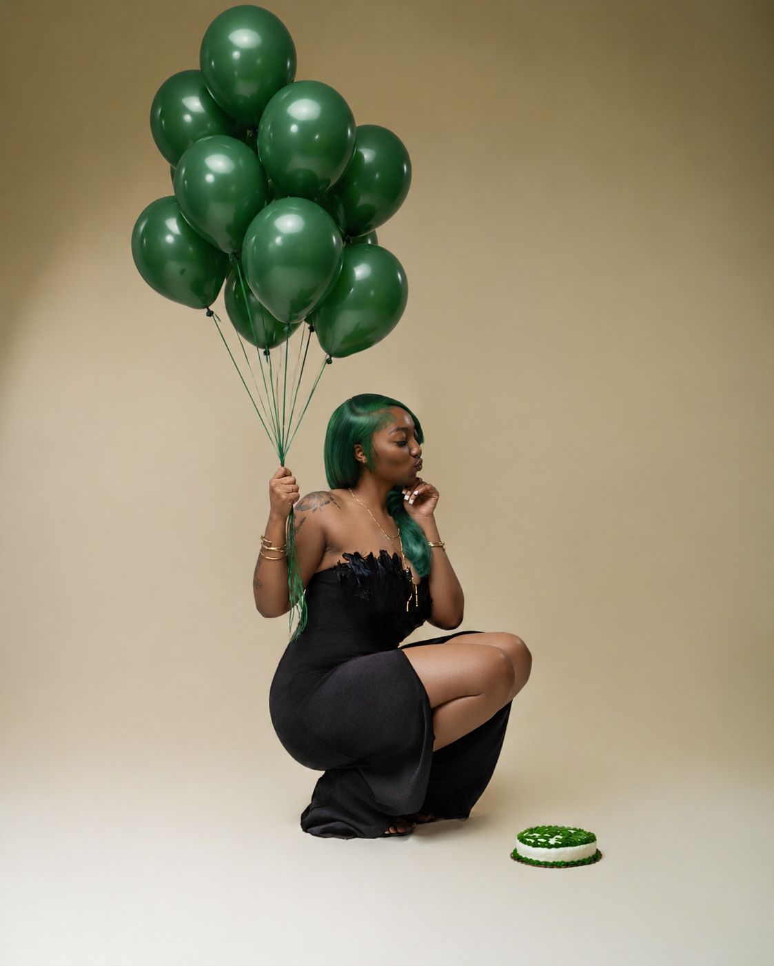 Woman in black dress holding green balloons beside a green cake, sitting gracefully in a studio setting.