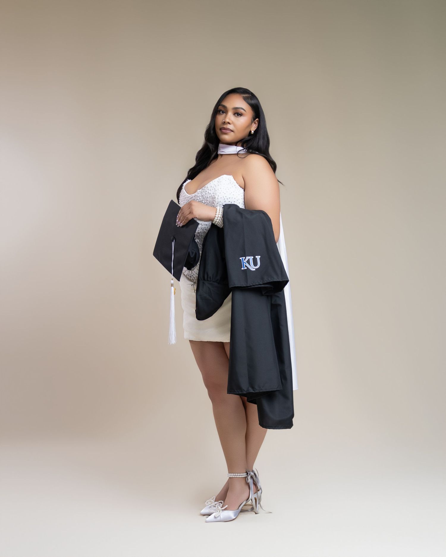 A woman poses in a white strapless dress holding a black KU graduation gown, wearing silver heels.