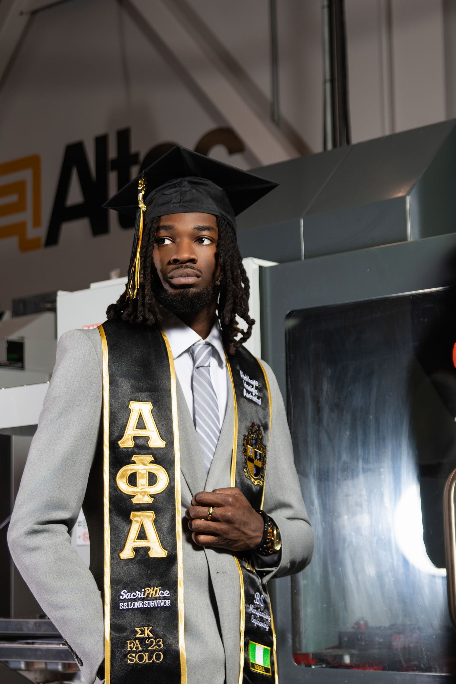 Graduate in cap and Alpha Phi Alpha stole poses proudly in industrial setting wearing suit and tie.