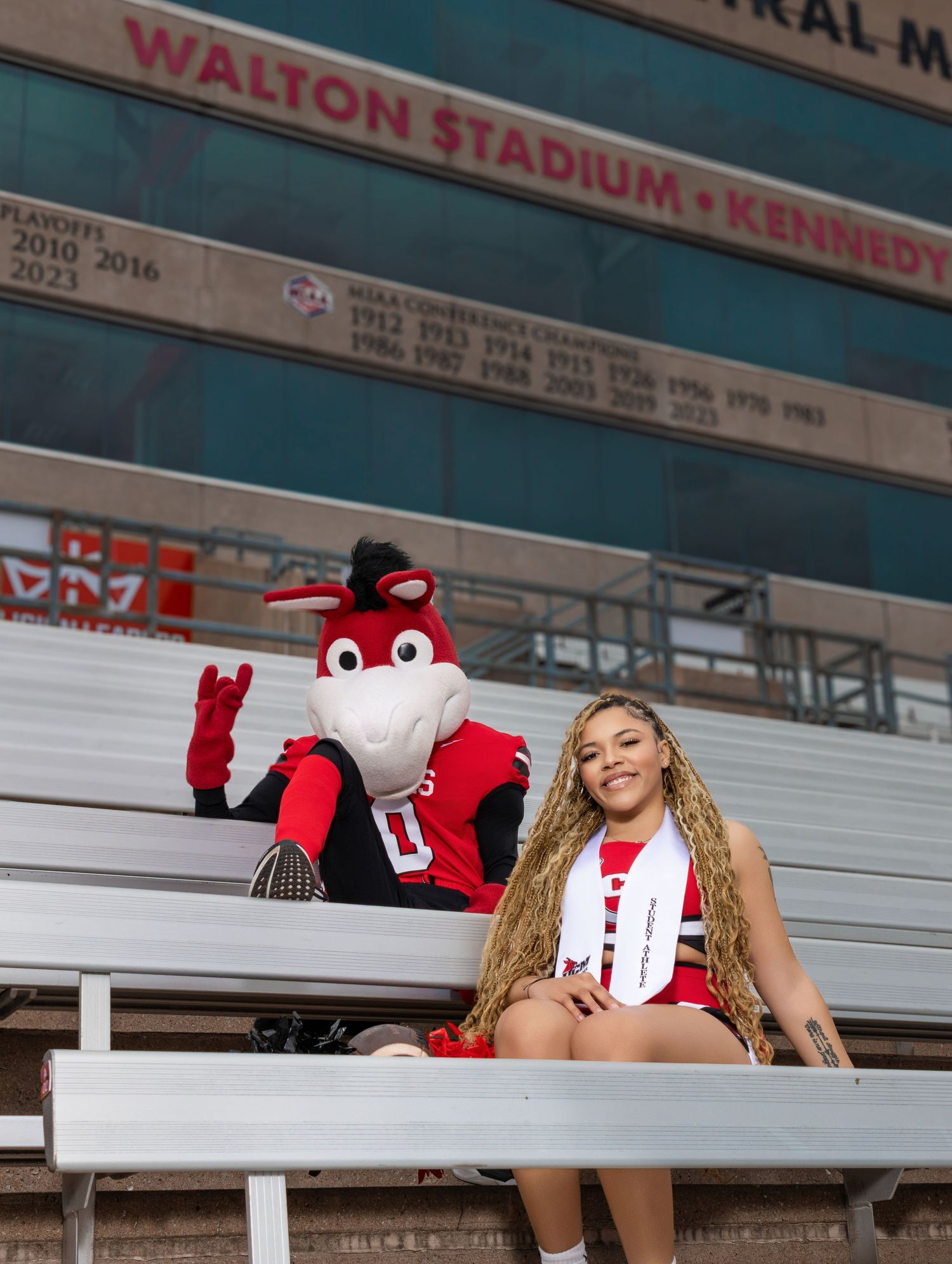 A cheerleader poses with a bull mascot in stadium bleachers at Walton Stadium.