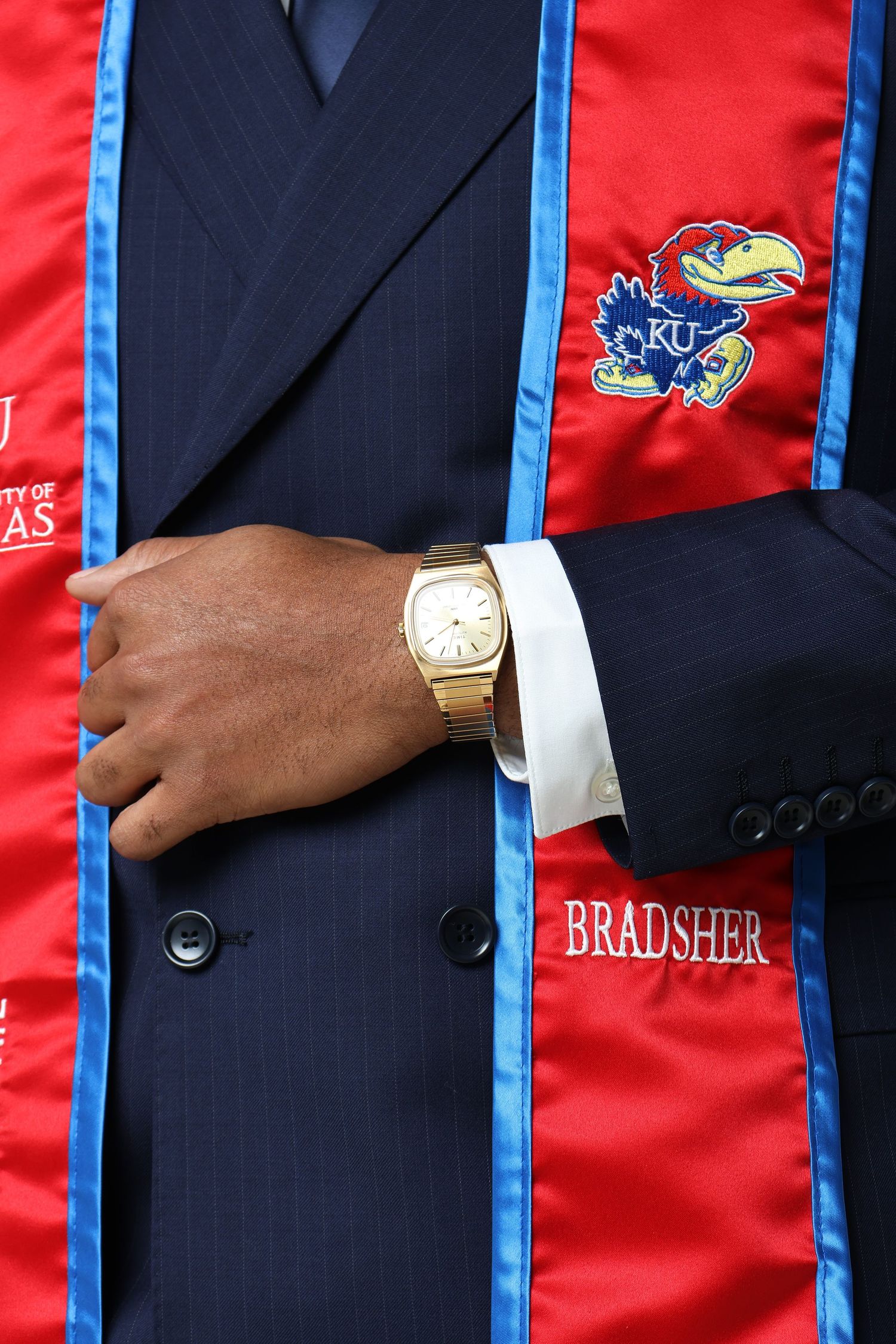 University of Kansas KU graduation stole worn over navy suit with gold watch, name BRADSHER embroidered on sash.