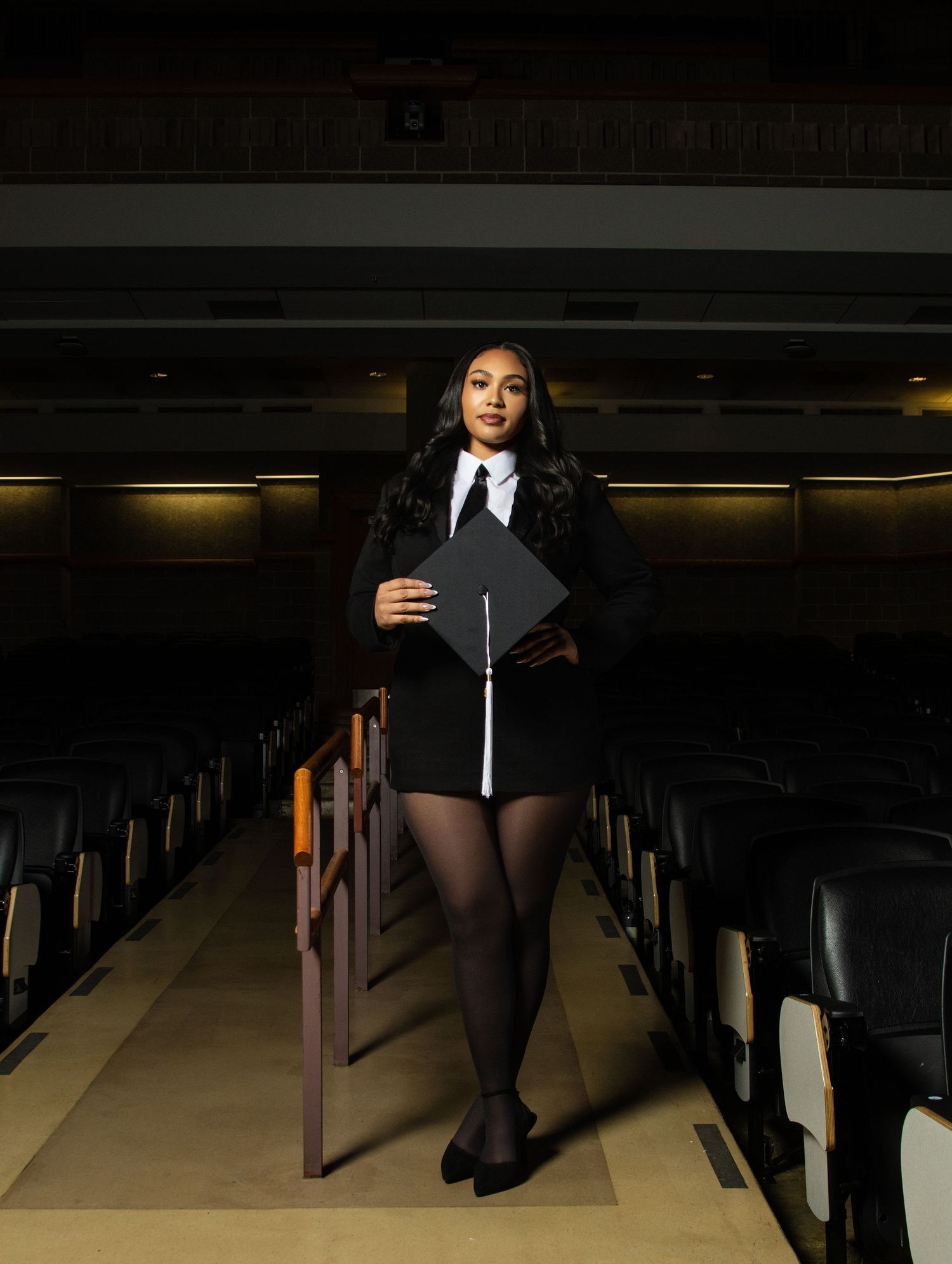 Graduate in black cap and gown poses proudly in an auditorium, holding her graduation cap.