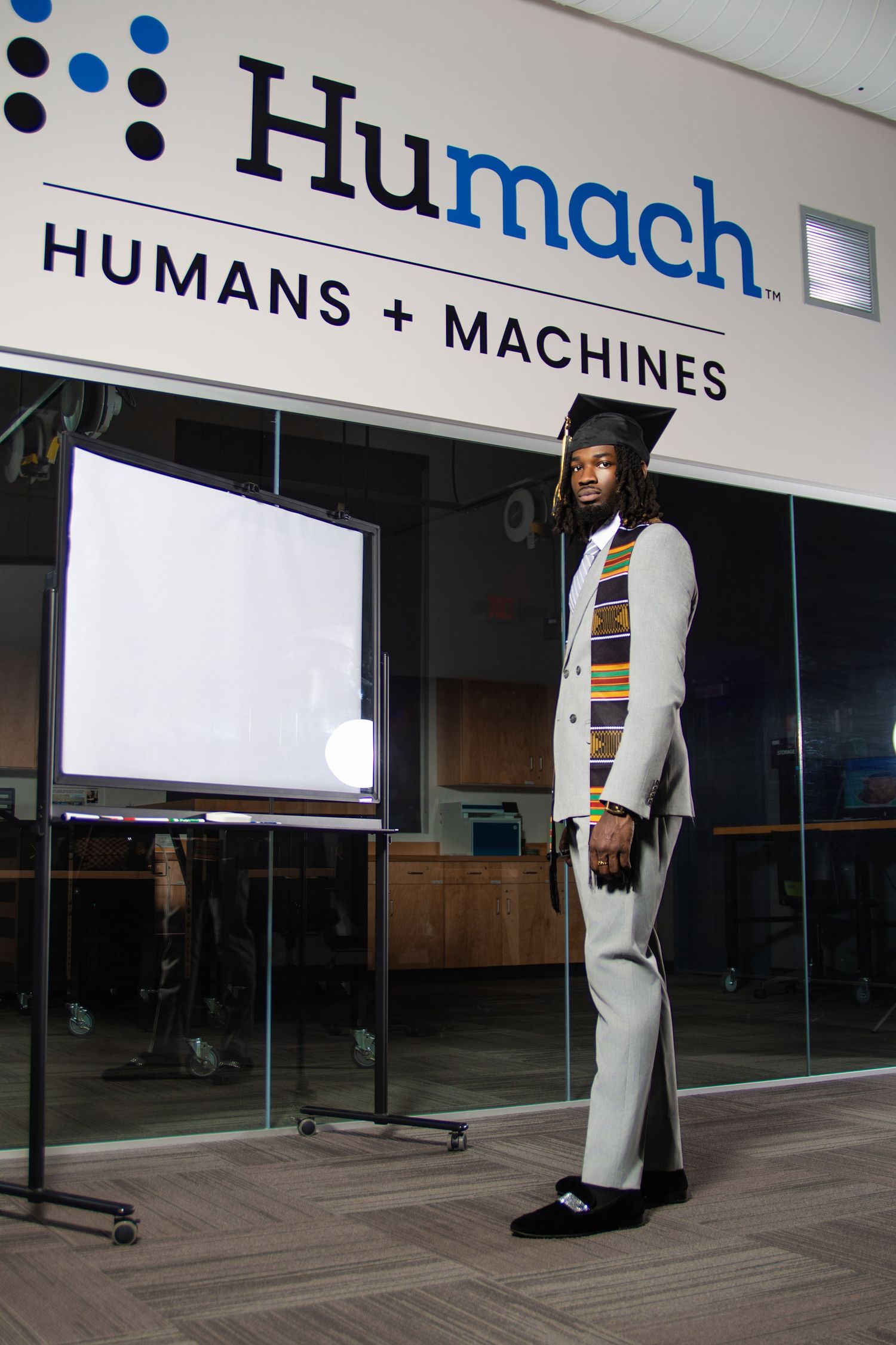 Graduate in cap and gown with kente stole stands proudly in front of Humach Humans + Machines office sign.
