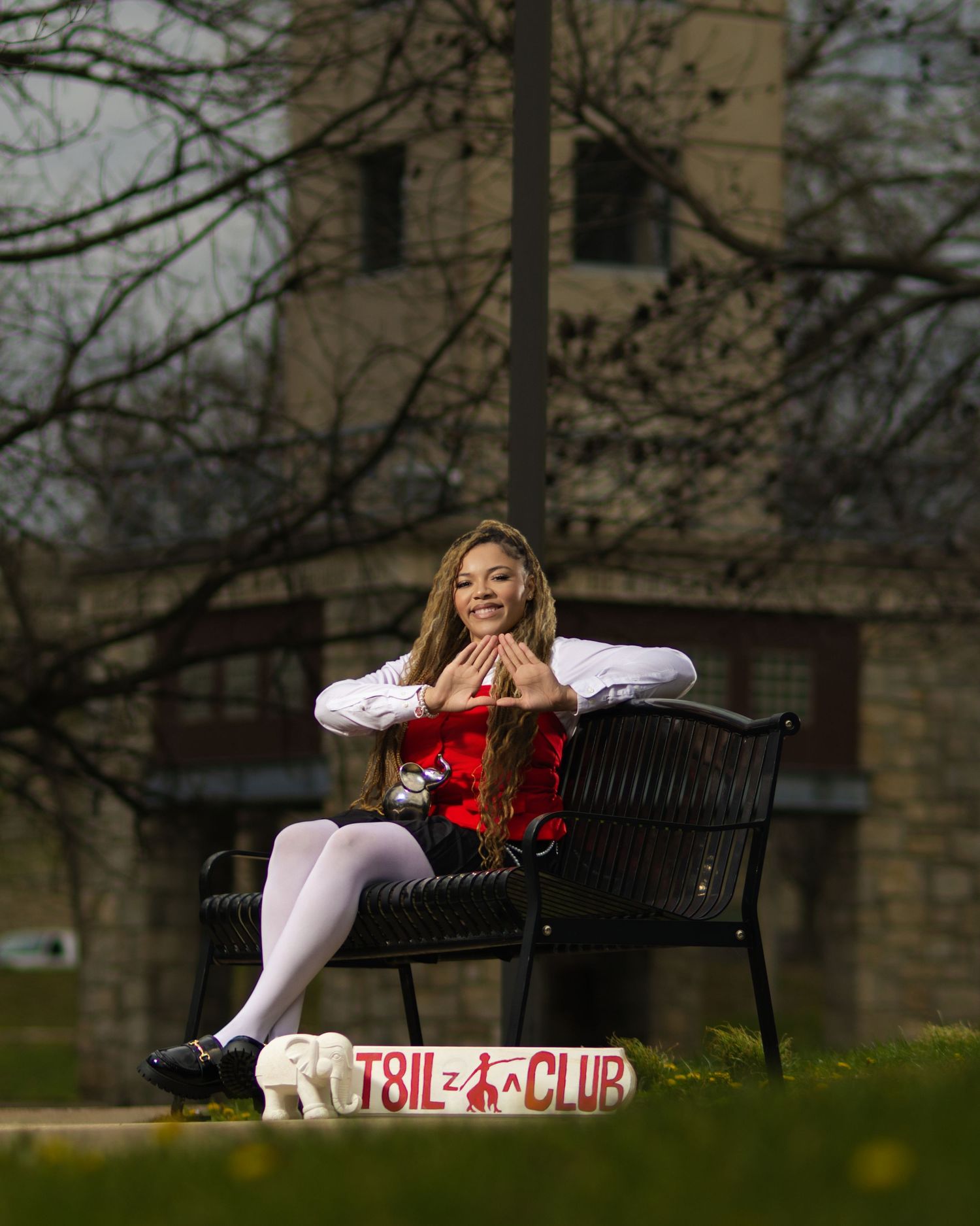 A young woman in a red and white outfit sits on a bench outdoors, posing with a T8IL Club sign nearby.