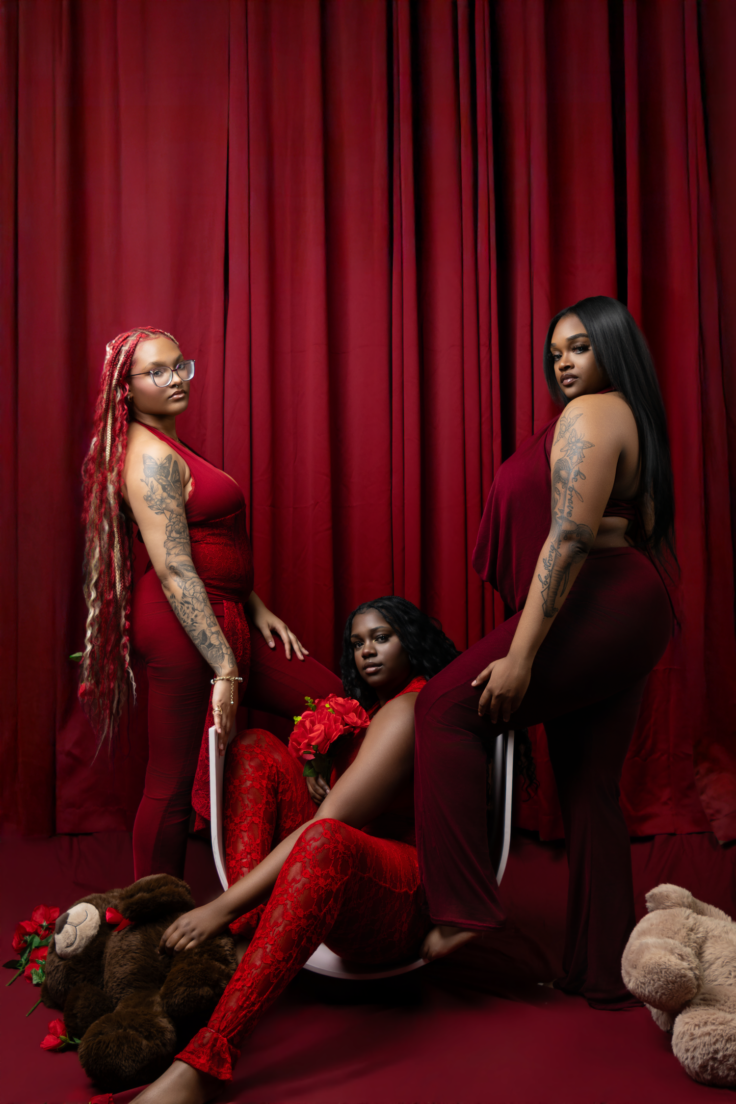 Three women in red outfits pose dramatically against a red curtain backdrop with roses and teddy bears.