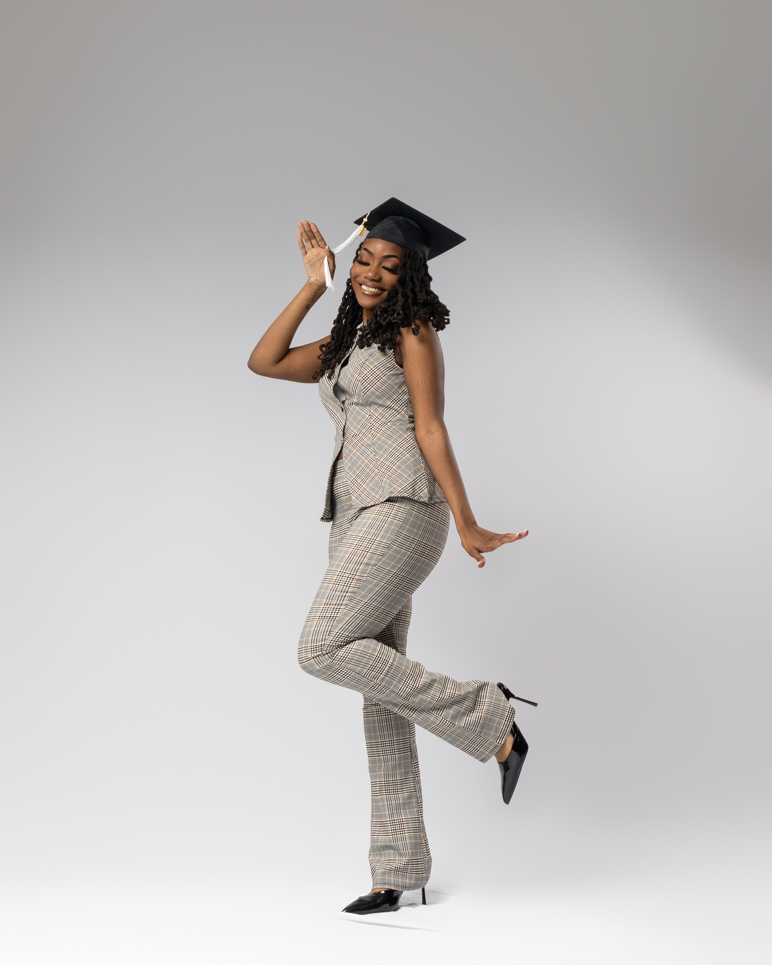 A young Black woman in a plaid pantsuit and graduation cap poses joyfully on one leg against a white background.