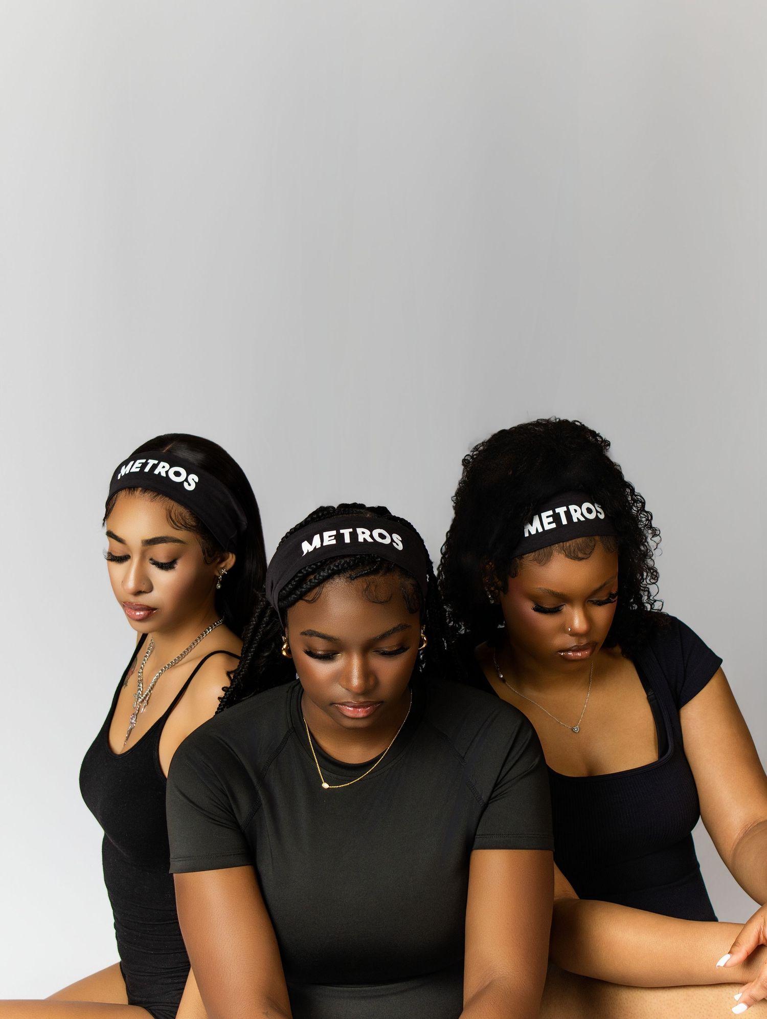 Three women wearing black METROS headbands and outfits pose together against a light gray background.