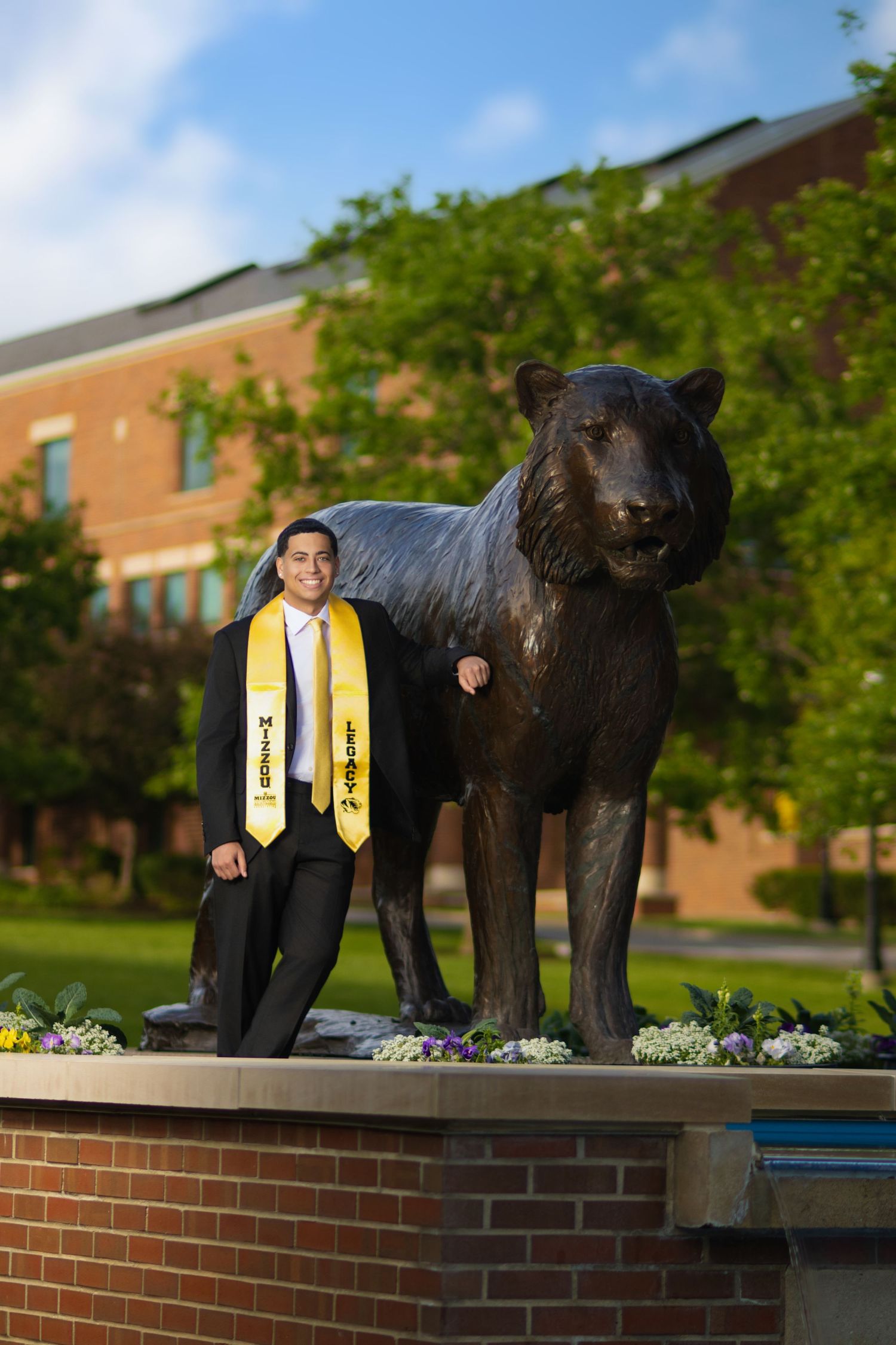 Graduate in black suit and gold stole poses beside a bronze tiger statue on a brick pedestal at a university campus.