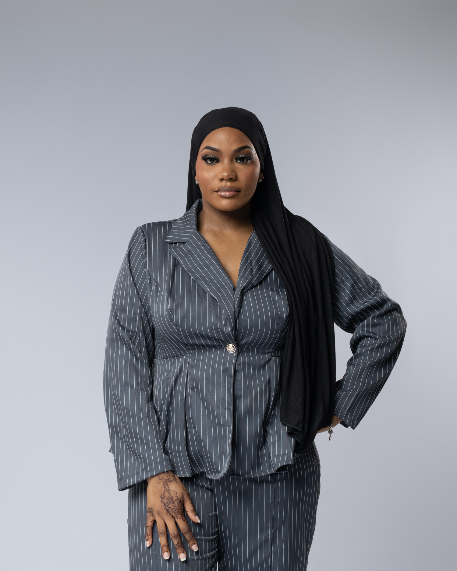 Woman in gray pinstripe blazer and matching pants with black hijab poses confidently against light gray background.
