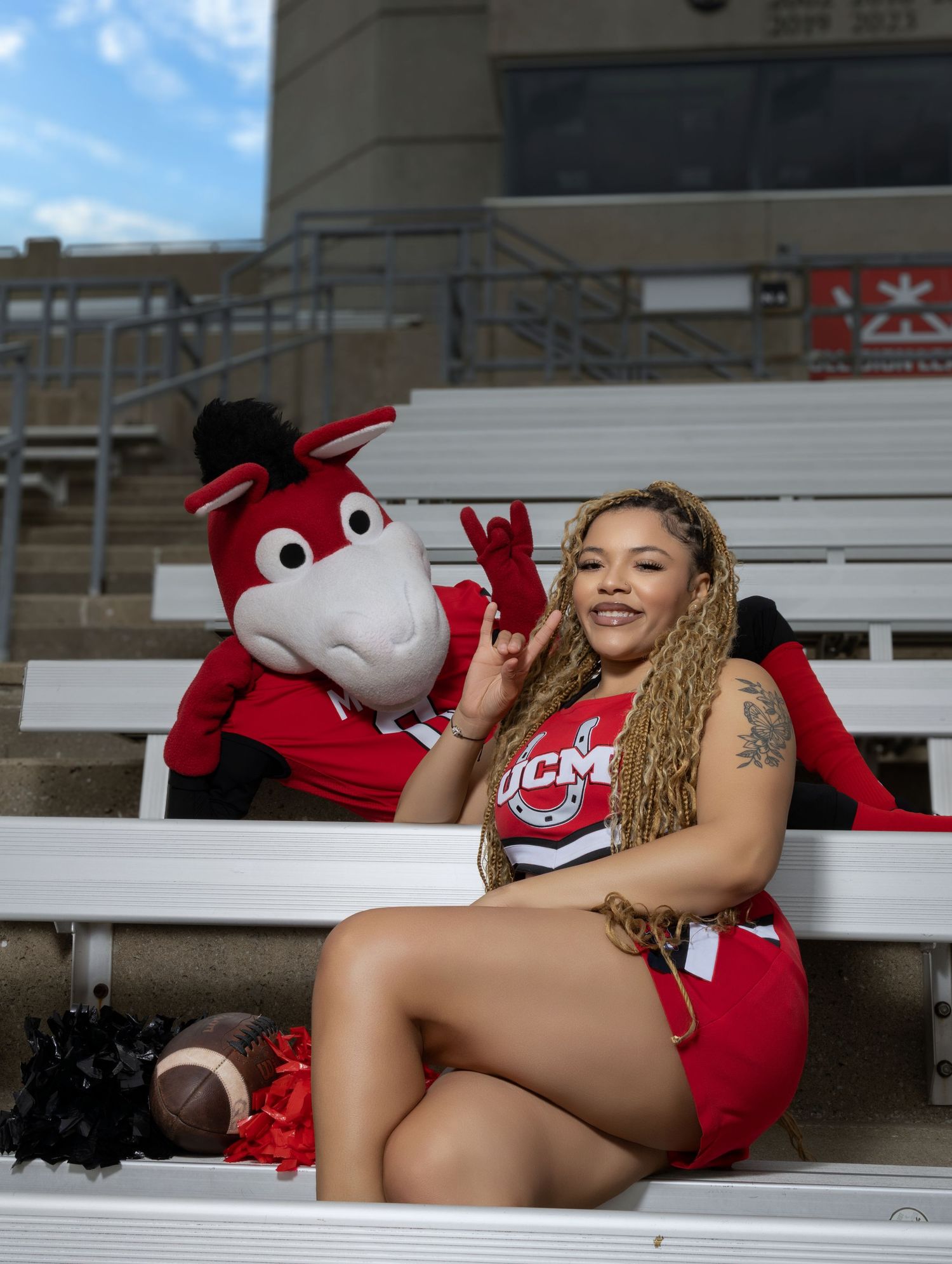 UCM cheerleader poses with red bull mascot in stadium bleachers, flashing peace signs in red and white uniform.