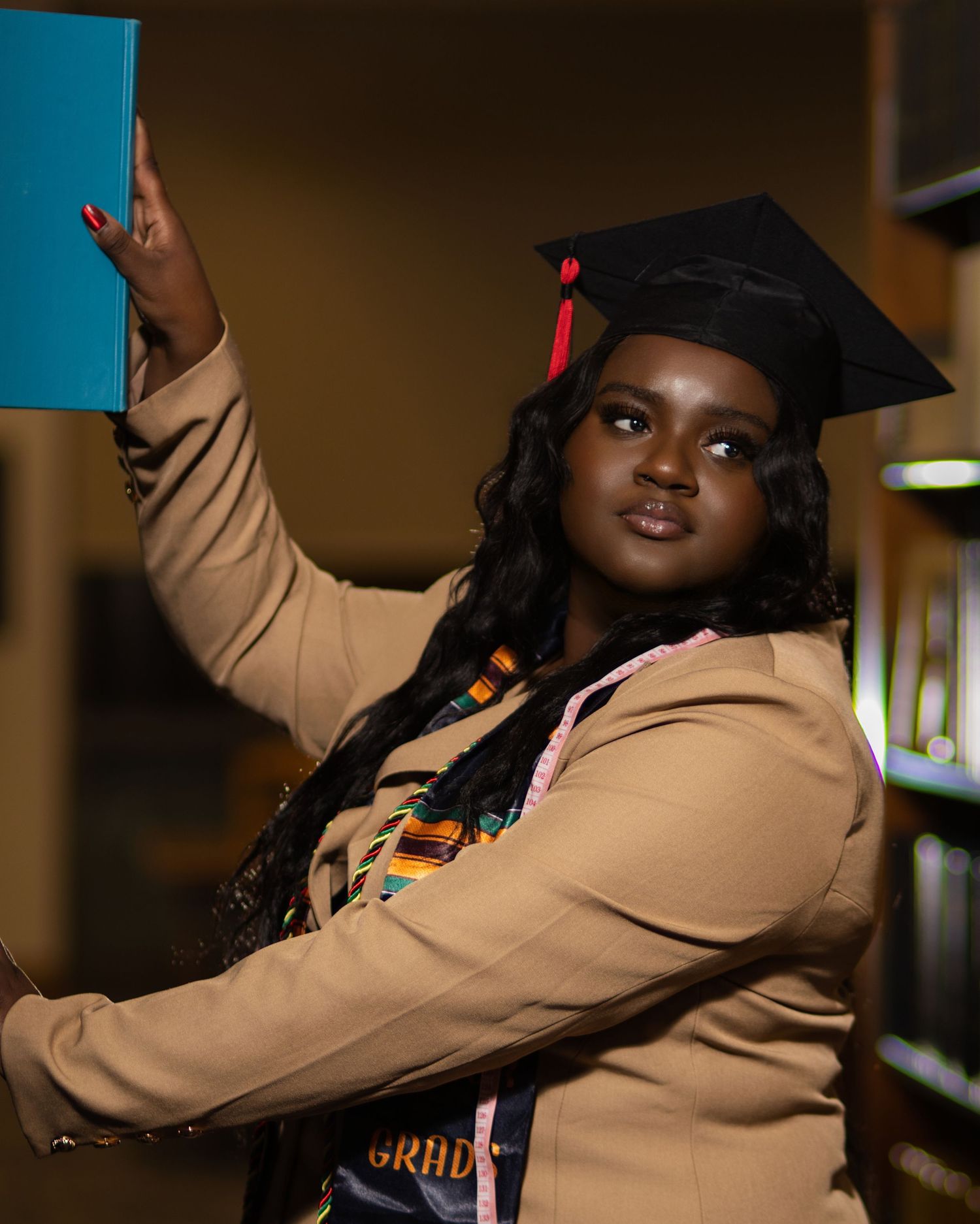 A young woman in graduation cap and tan blazer holds a teal book aloft in a library setting.