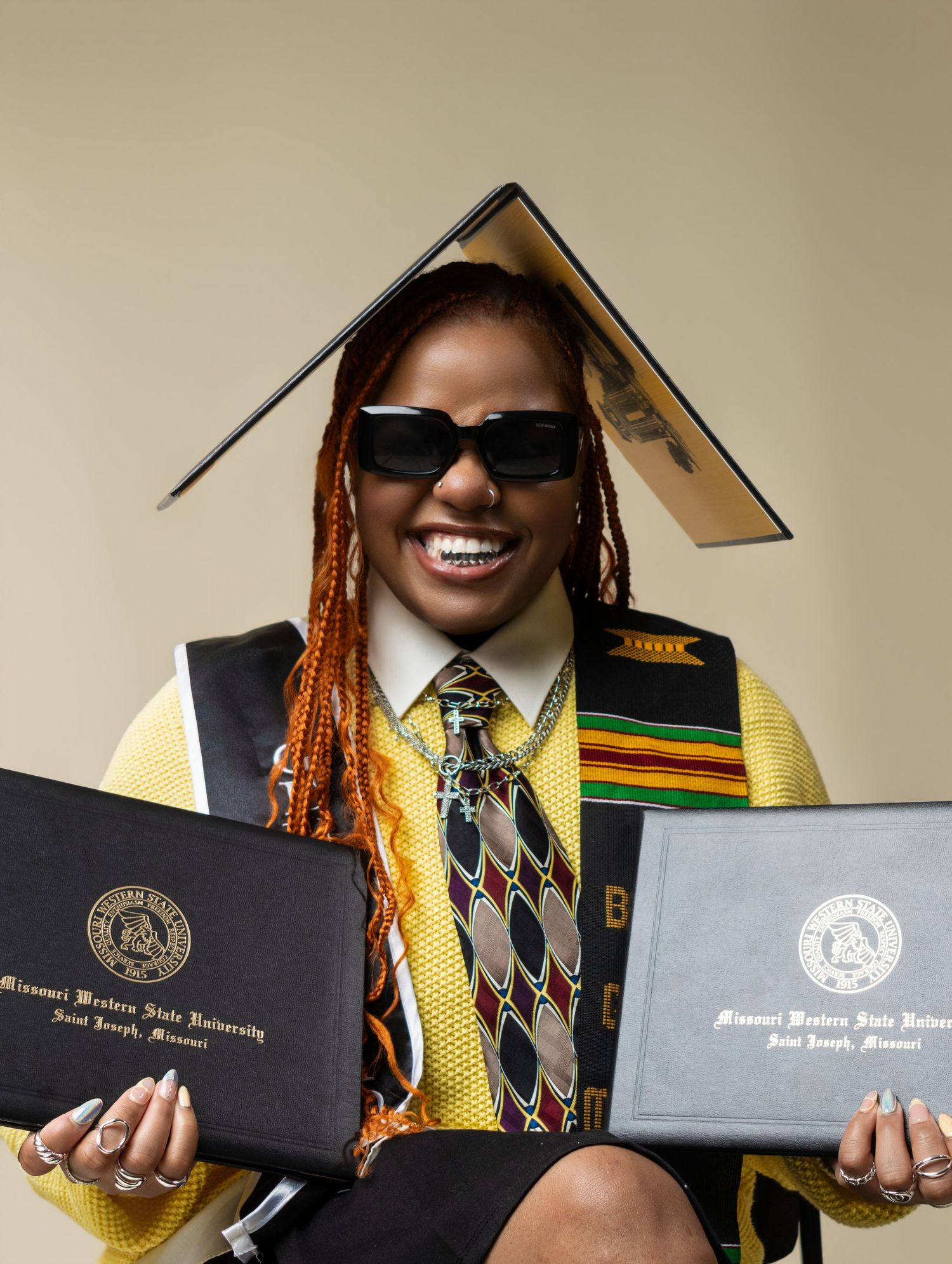 Graduate smiling in cap and gown holding two Missouri Western State University diplomas, wearing sunglasses and kente cloth stole.