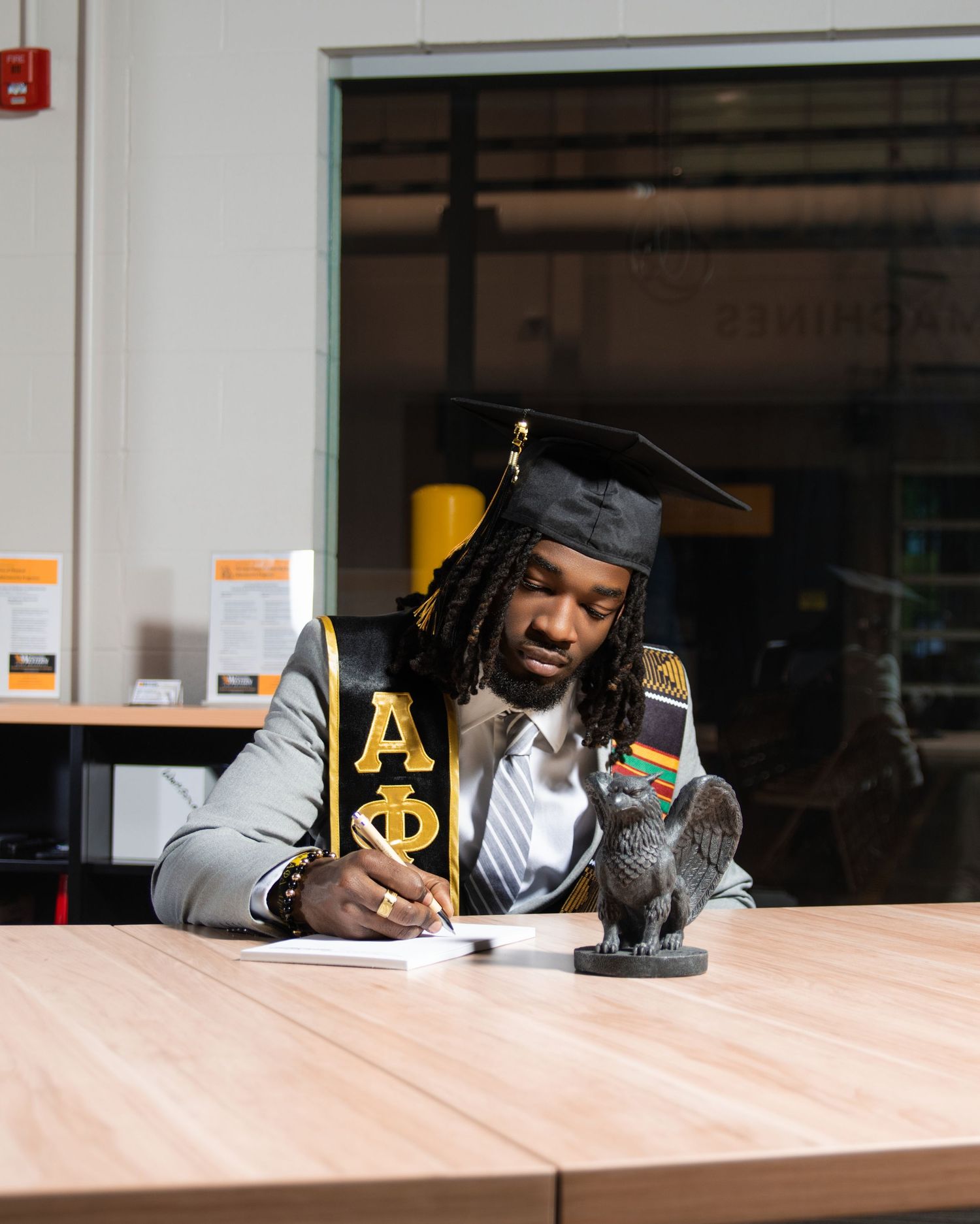 Graduate in cap and Alpha Phi sash signs document beside a small eagle statue on a wooden table.