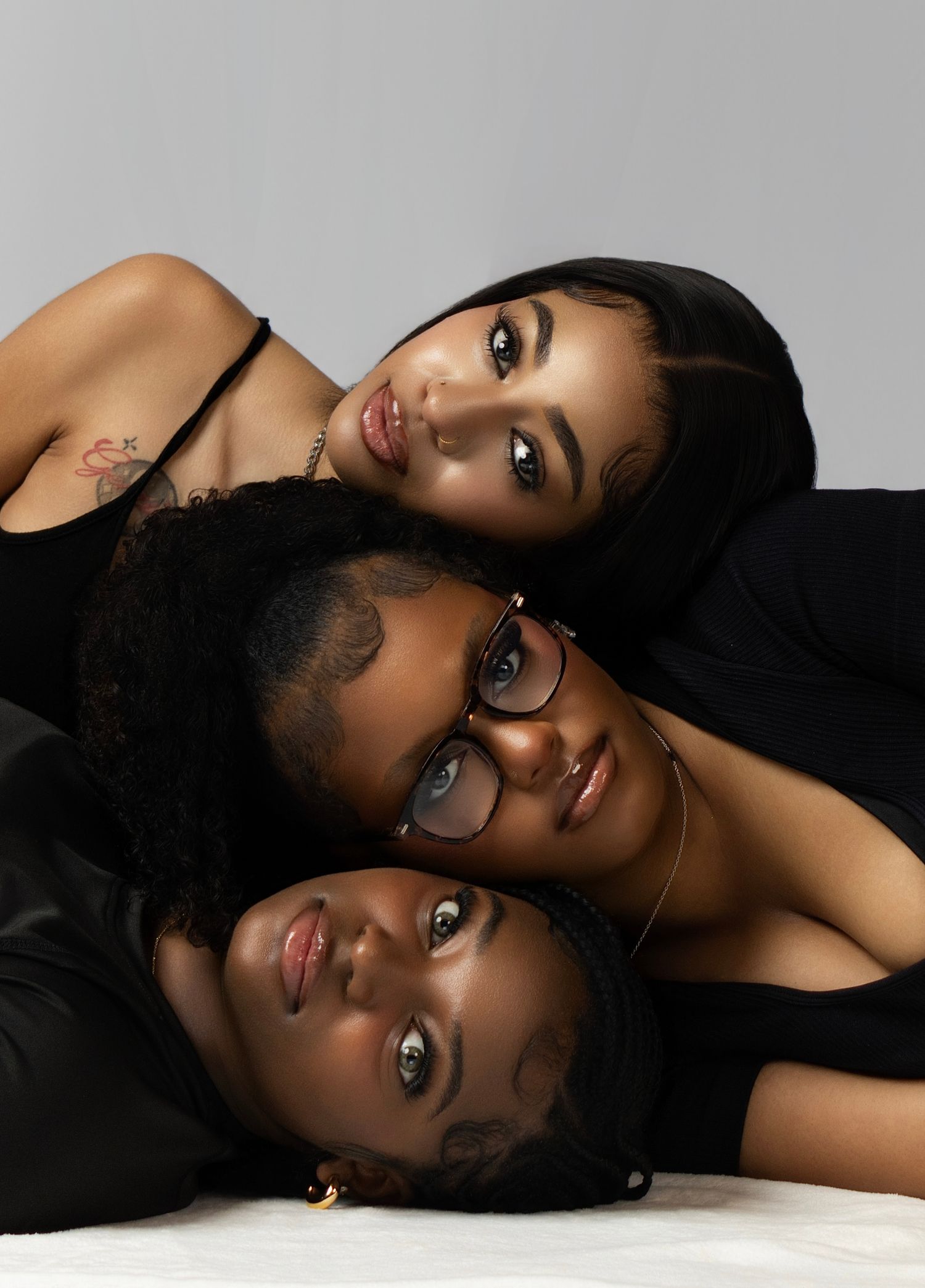Three young women in black outfits pose together in a studio portrait, stacked closely with soft gray background.