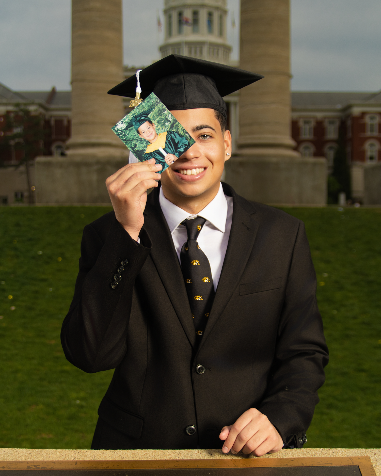 Smiling graduate in cap and gown holds a small photo card, posing outdoors near a university building.