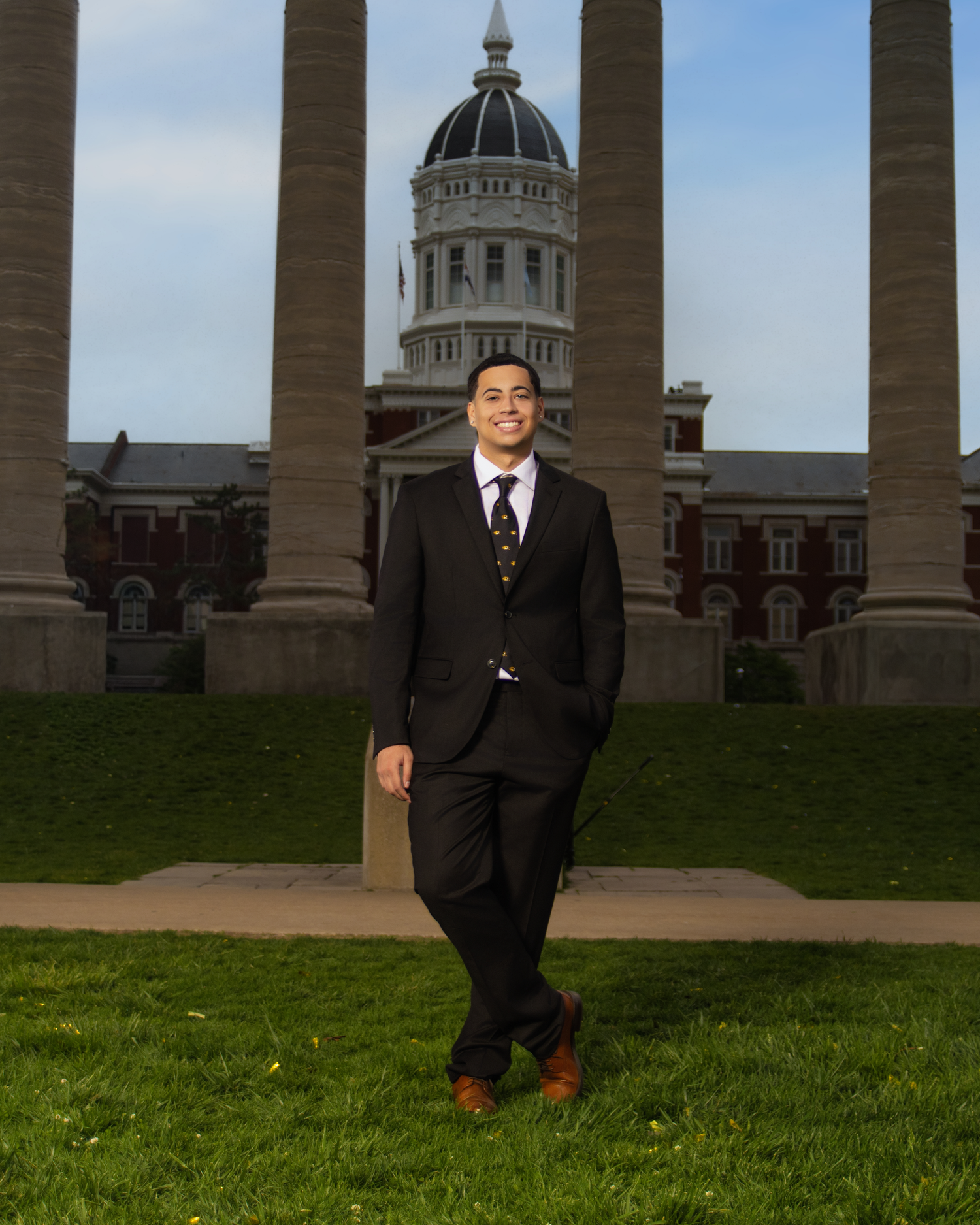 Young man in black suit stands on green lawn before historic domed building with tall columns.