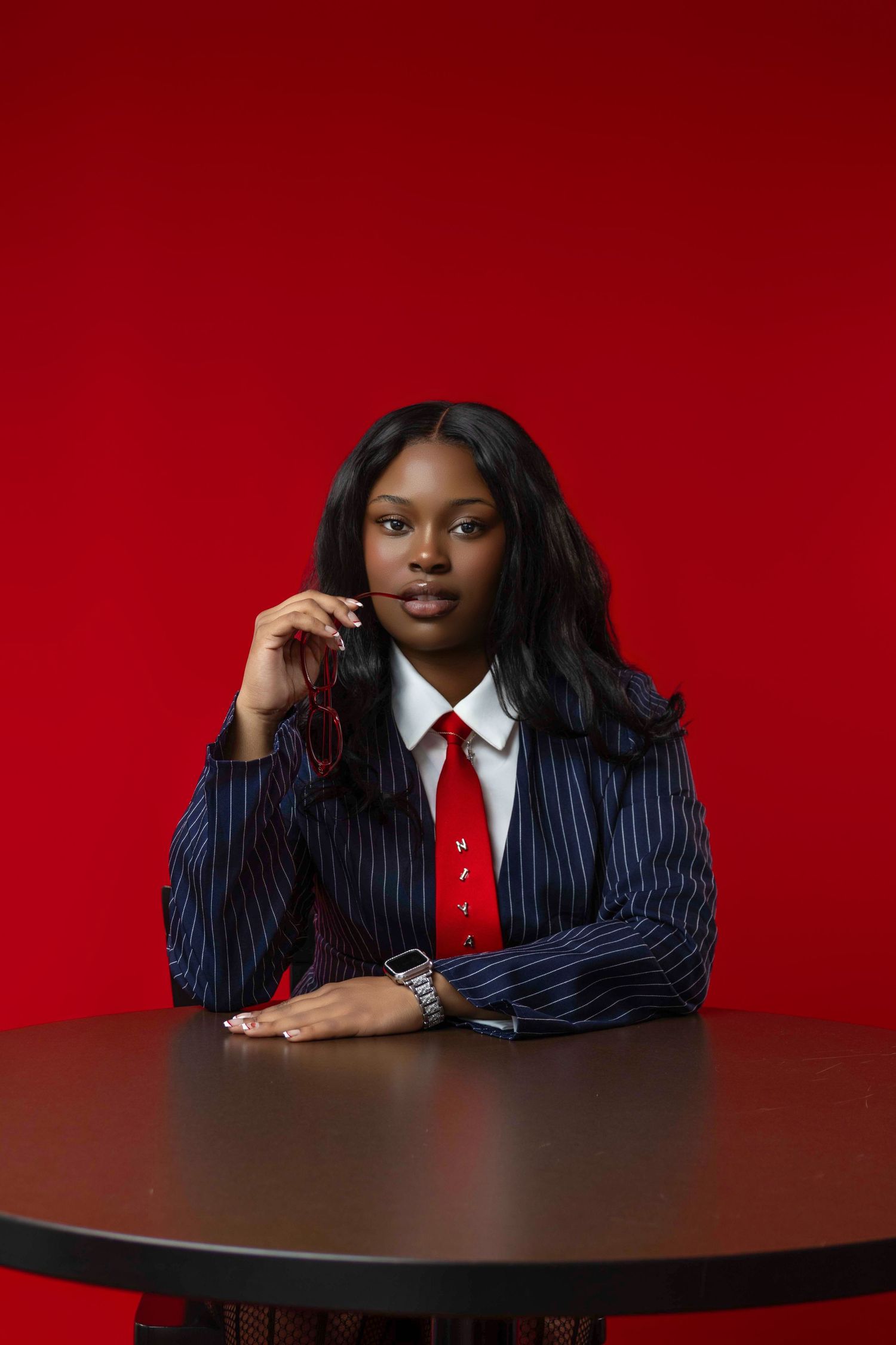 Young Black woman in pinstripe suit and red tie sits at dark table against bold red background.