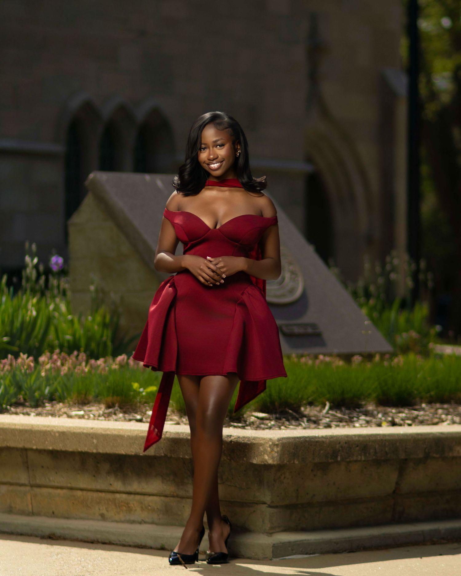 Woman in elegant red off-shoulder mini dress posing outdoors near stone architecture with lush green garden background.