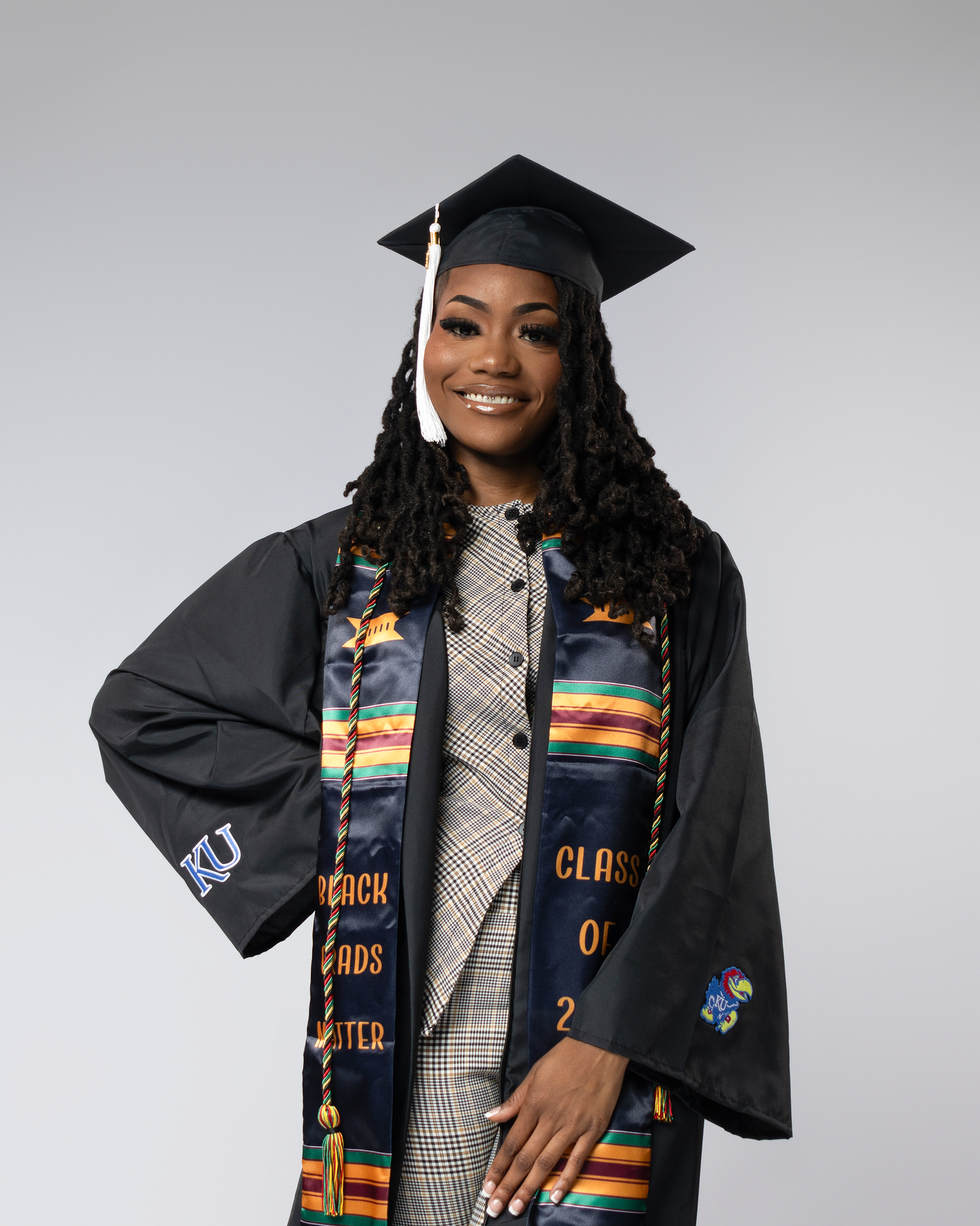 A smiling graduate wearing a black cap and gown with a colorful Kente stole at a University of Kansas graduation.