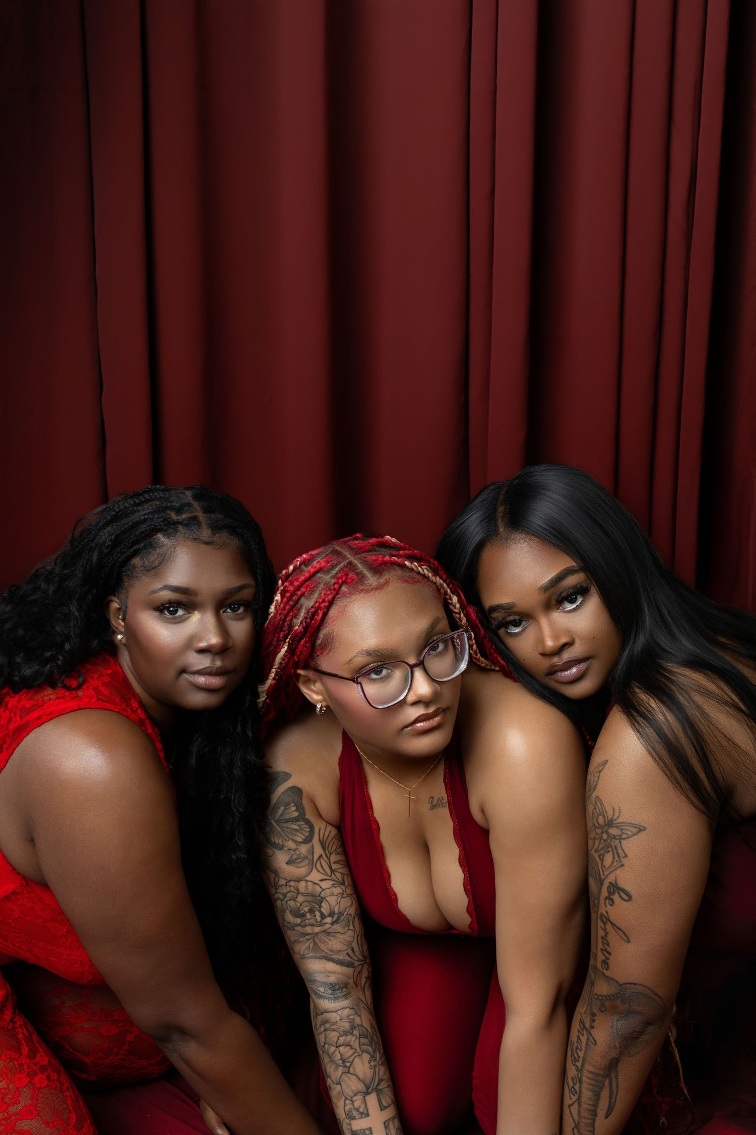 Three tattooed women in red outfits pose closely together against a dark red curtain backdrop.