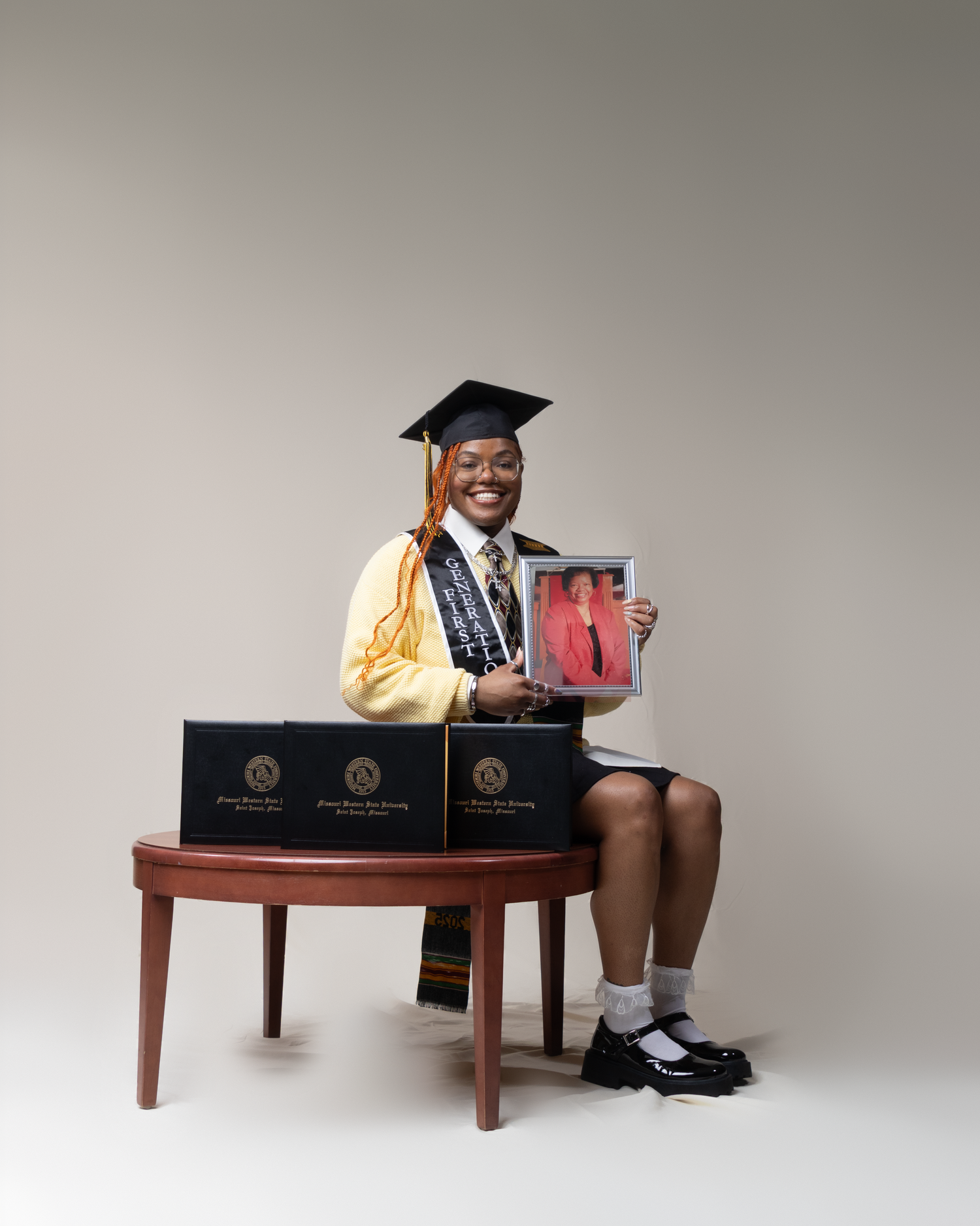Graduate in cap and gown sits on bench holding framed photo, with diploma boxes displayed beside them.