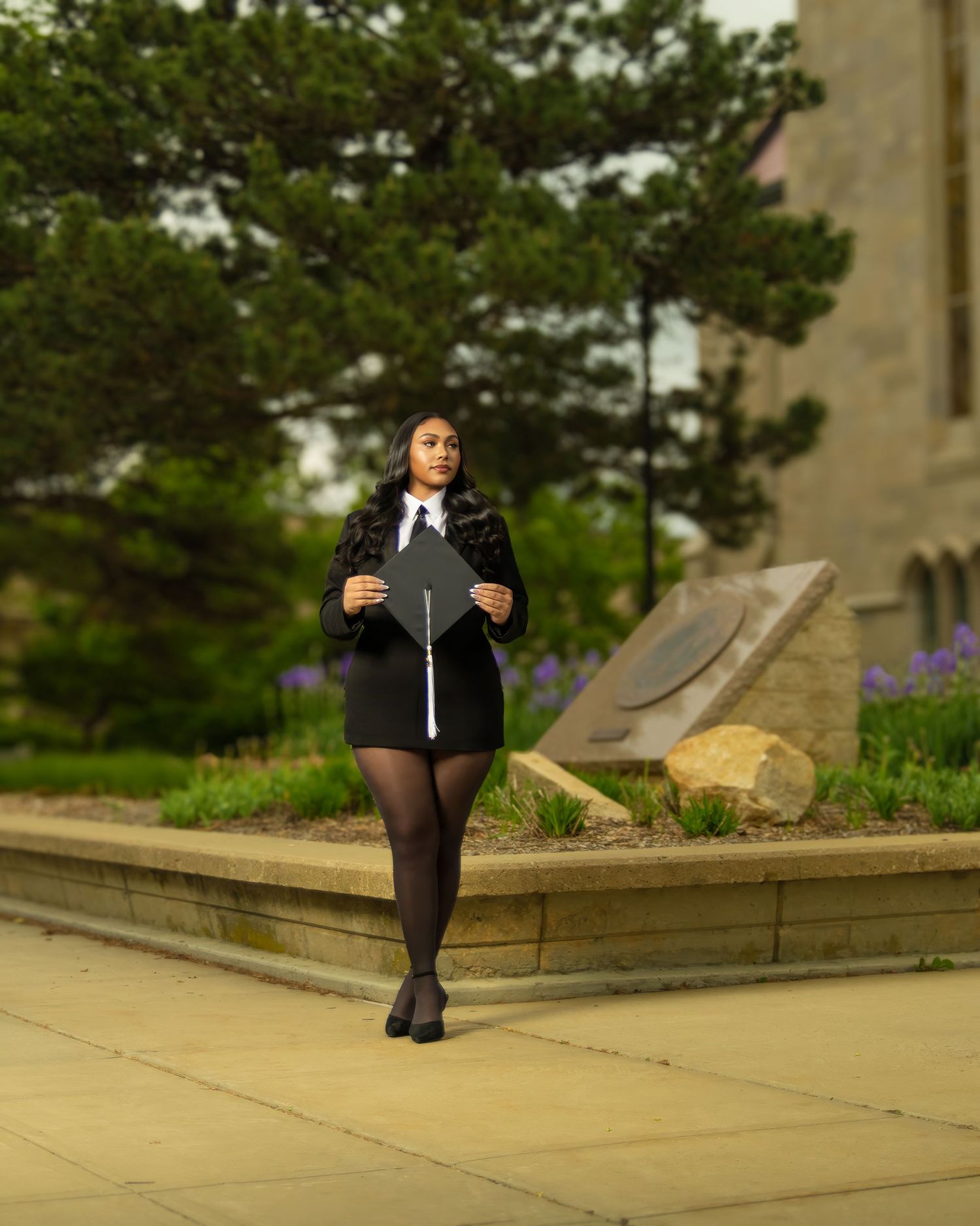 Graduate in black gown holds cap outdoors near stone building and lush greenery on college campus.