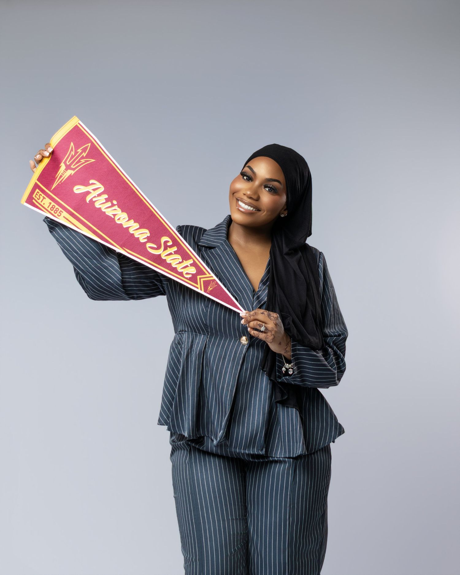 Woman in pinstripe suit smiling and holding an Arizona State University pennant flag on a gray background.