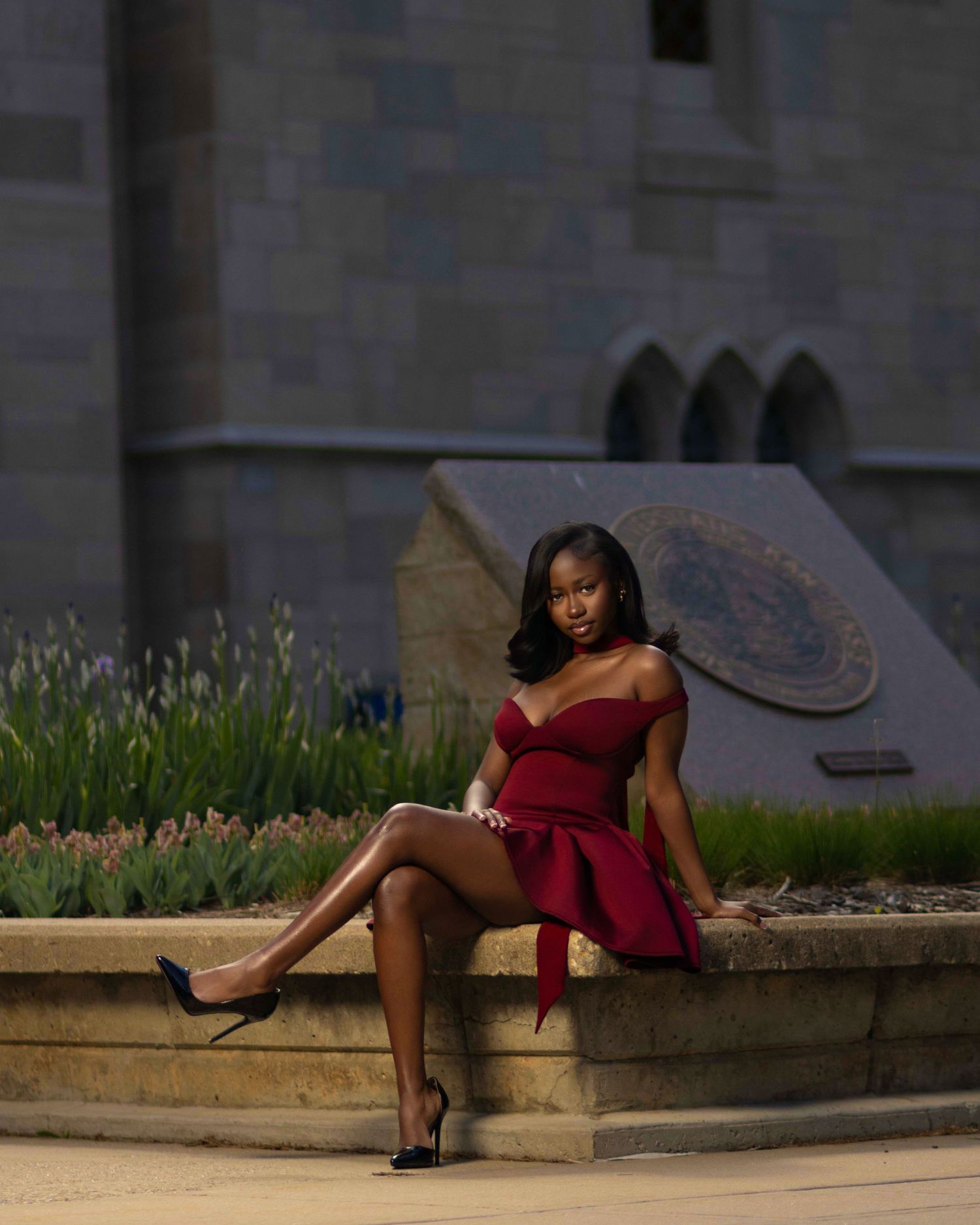 A young woman in a red off-shoulder mini dress poses gracefully on a stone ledge near a Gothic building.