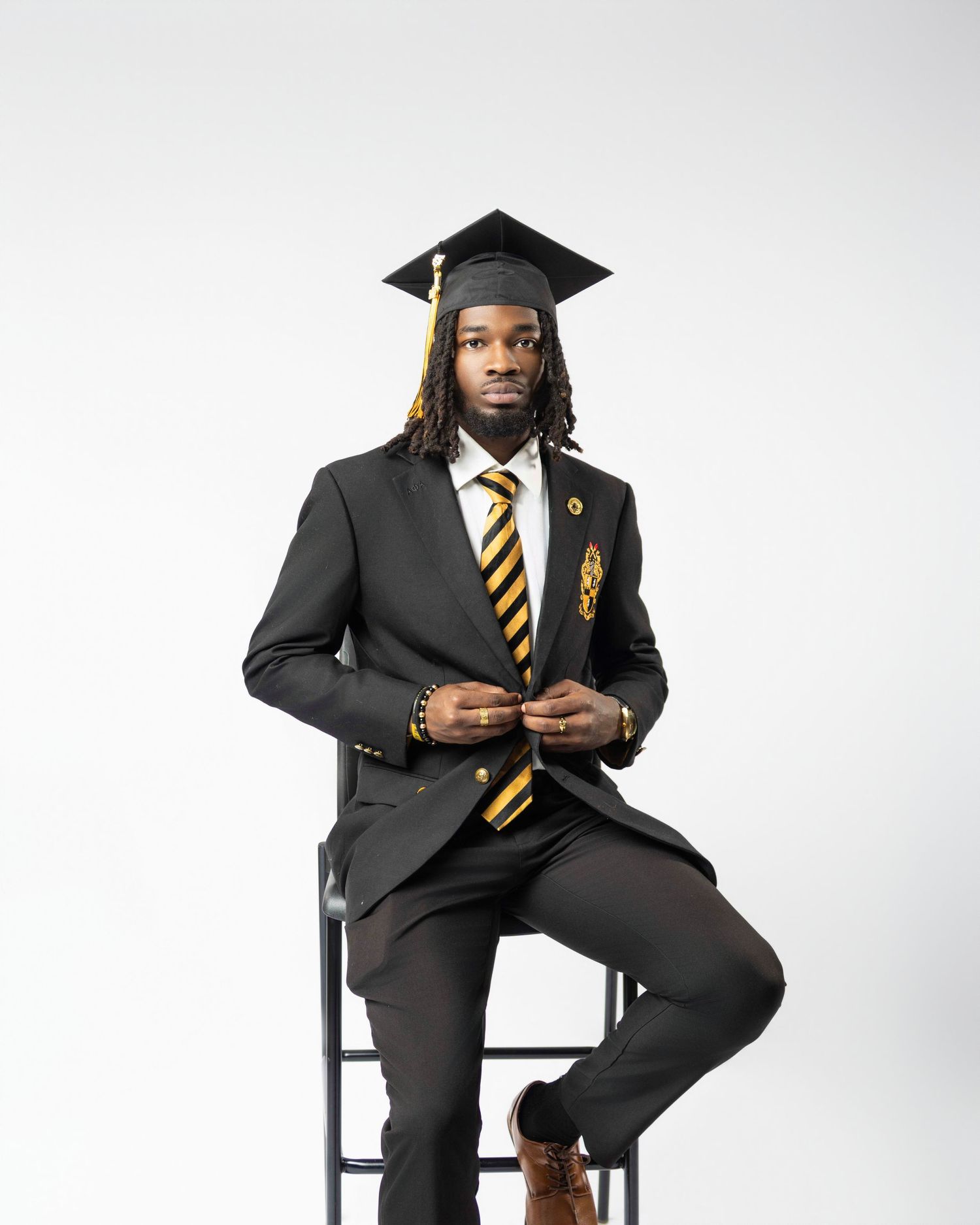 Young man in black suit and graduation cap poses confidently on stool in professional graduation photo.