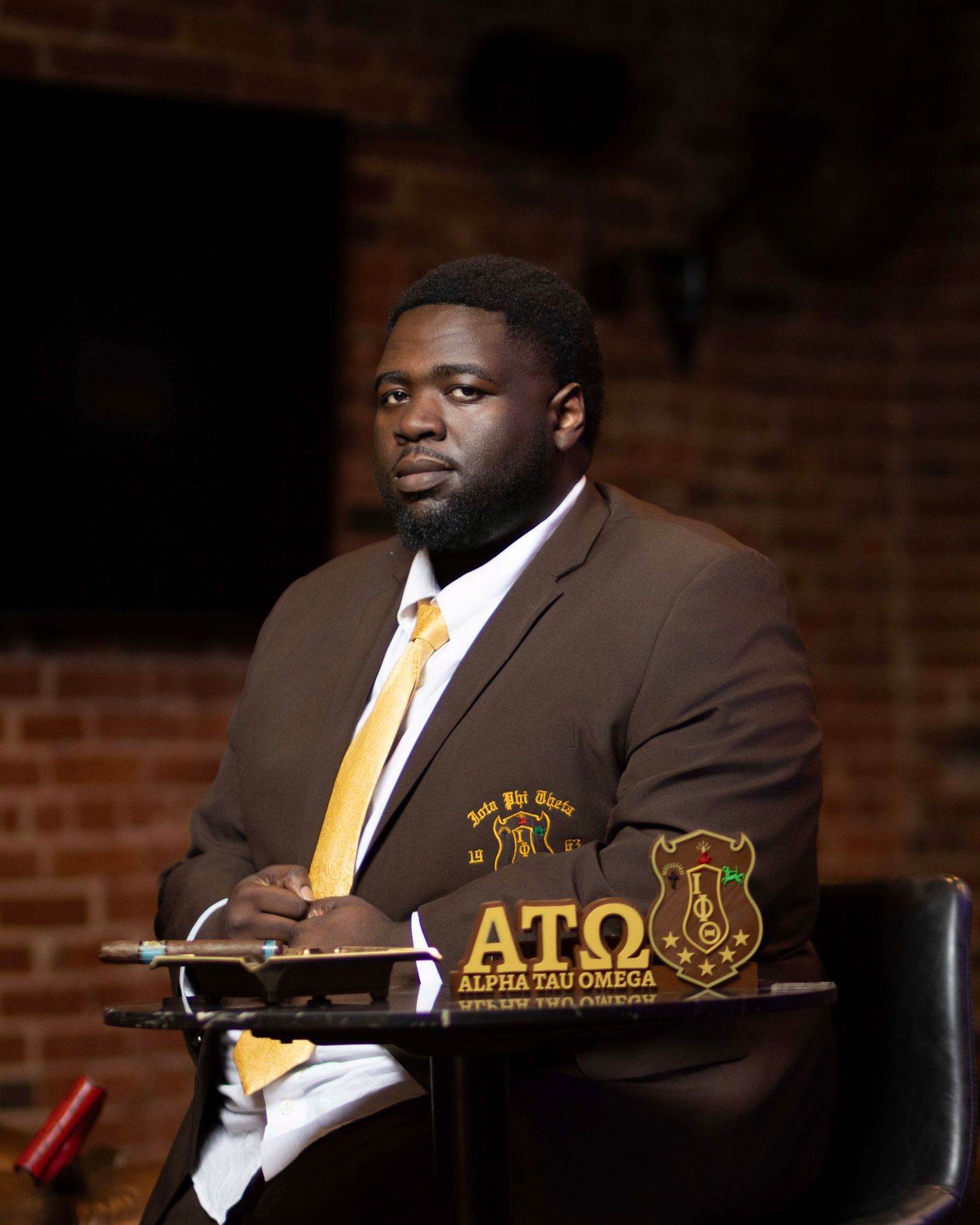 Man in Alpha Tau Omega brown blazer and gold tie sits formally at a podium nameplate.
