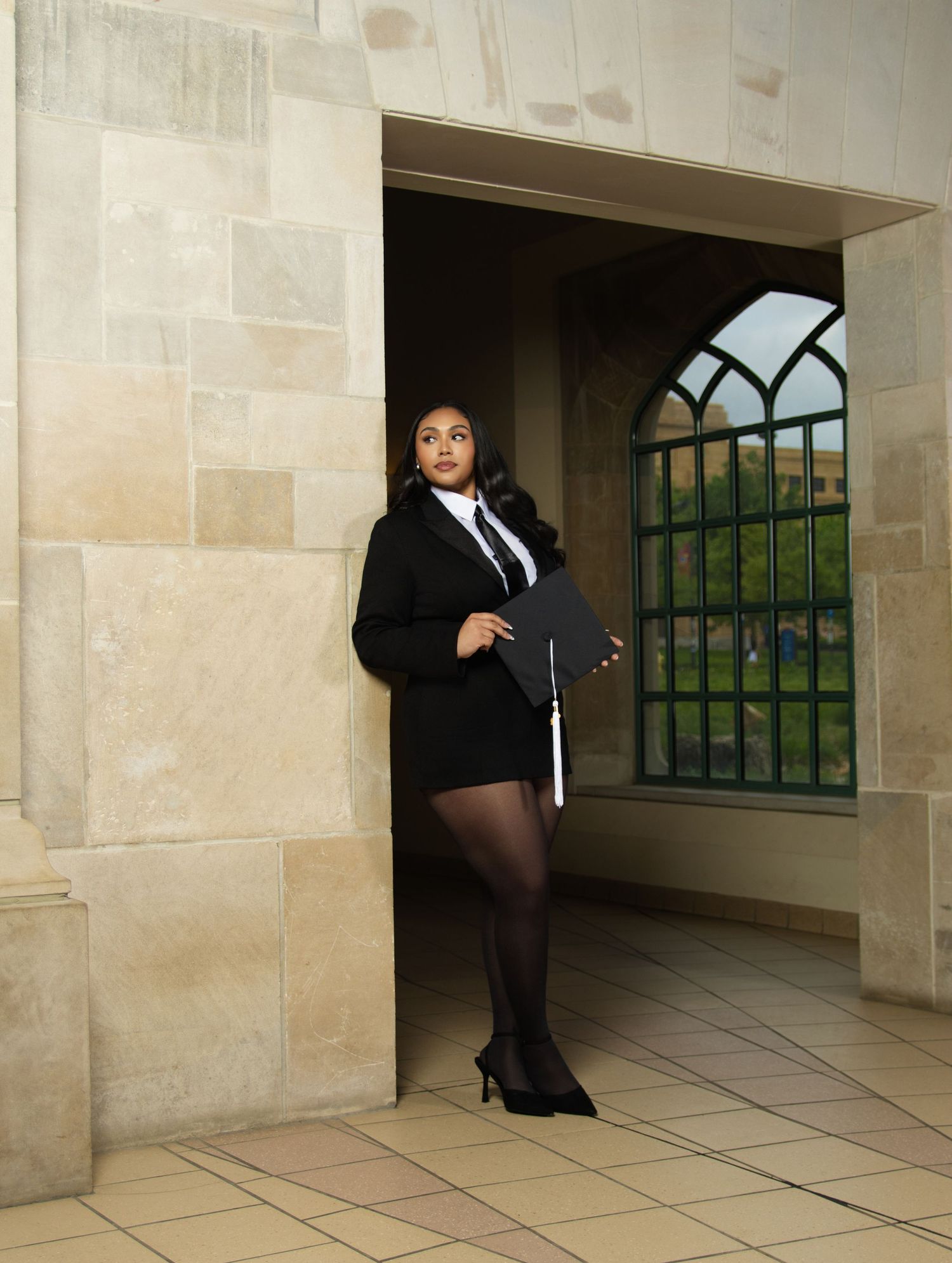 Graduate in black academic gown and mortarboard poses confidently in stone archway holding diploma.