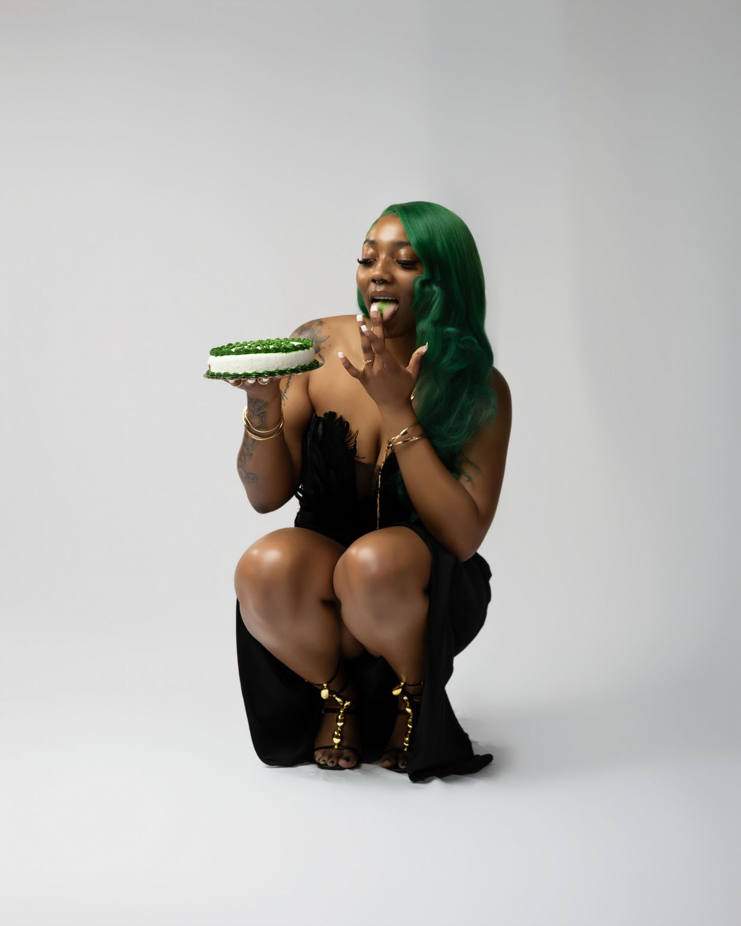 Woman with green hair crouching in black outfit holding a decorated cake, wearing gold accessories and boots.