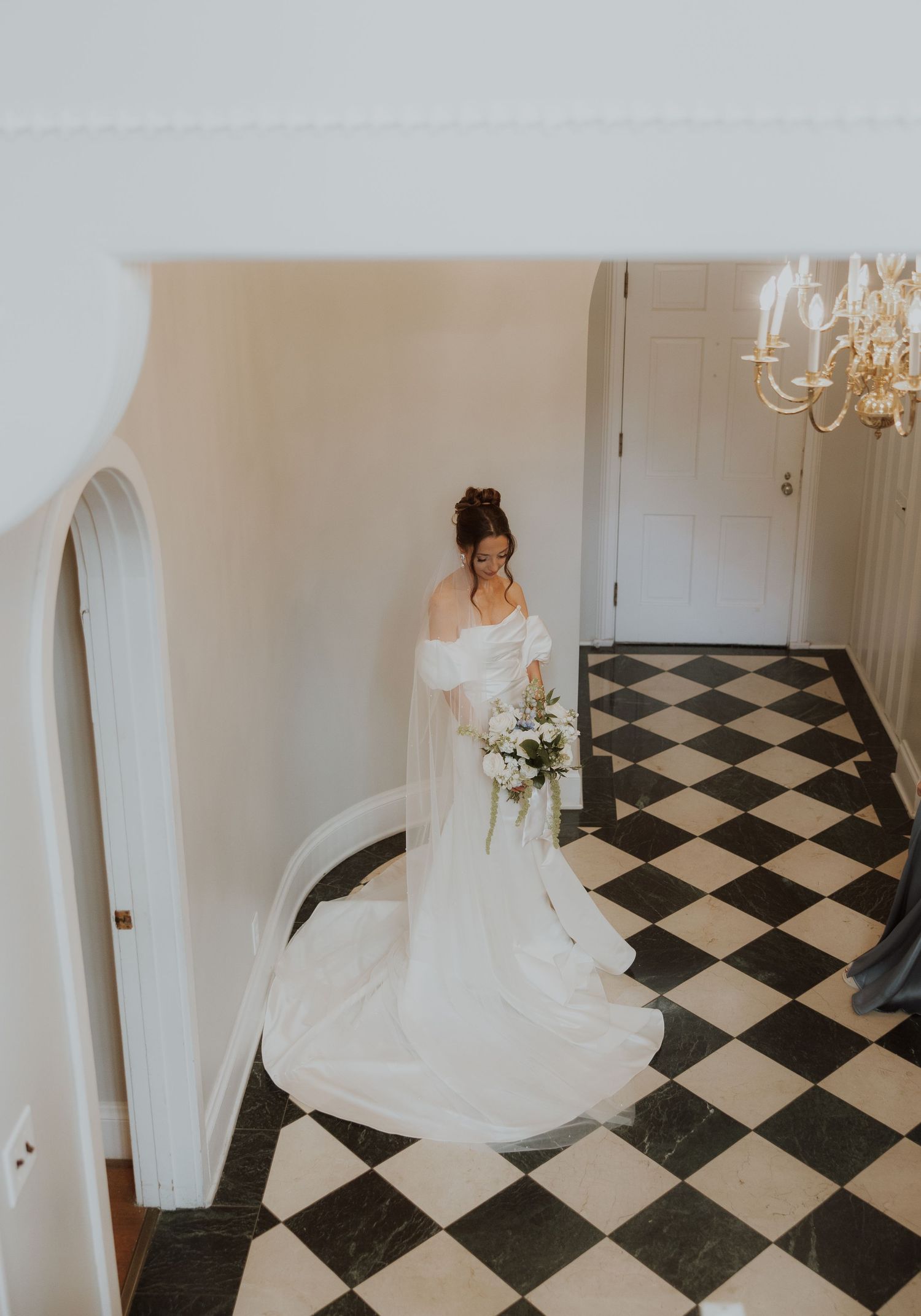 Bride in flowing white wedding dress stands on dramatic black and white checkered floor in elegant interior space.