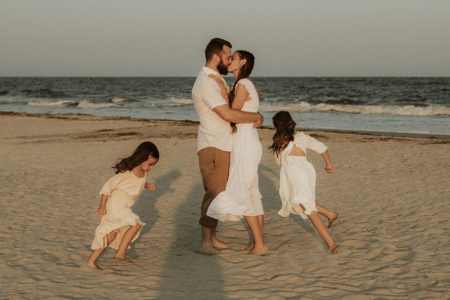 A family plays together on a sandy beach at sunset with children jumping and parents embracing.