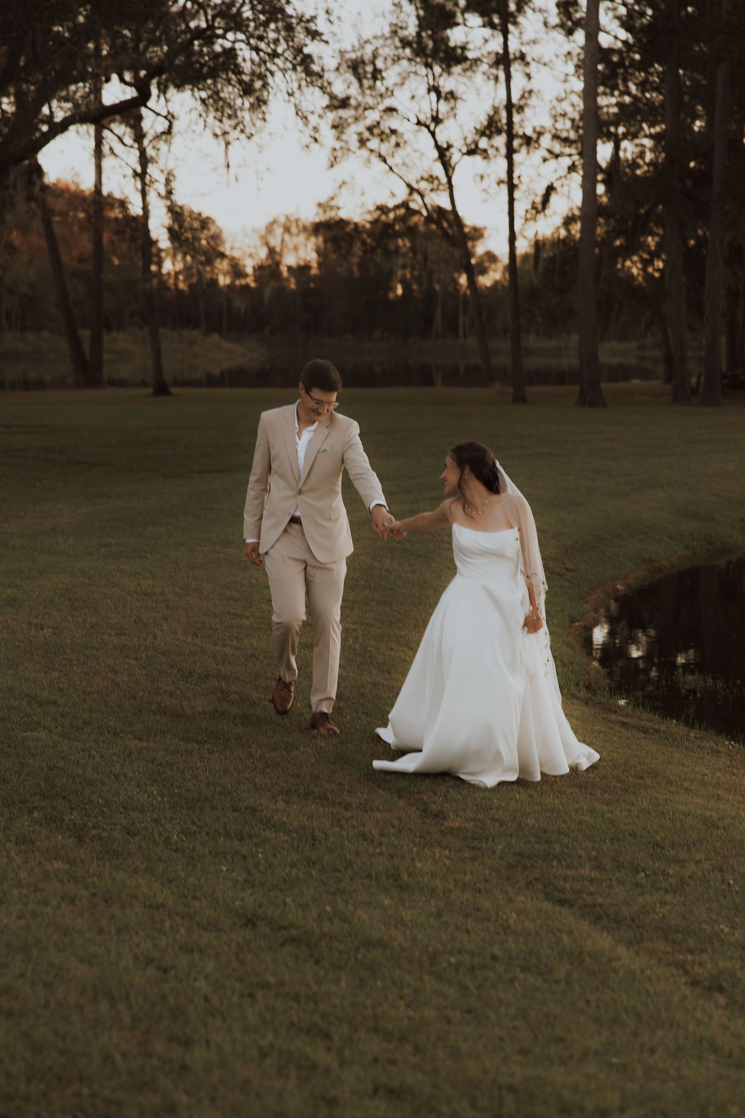 A couple walks hand in hand across a sunset-lit lawn in wedding attire.