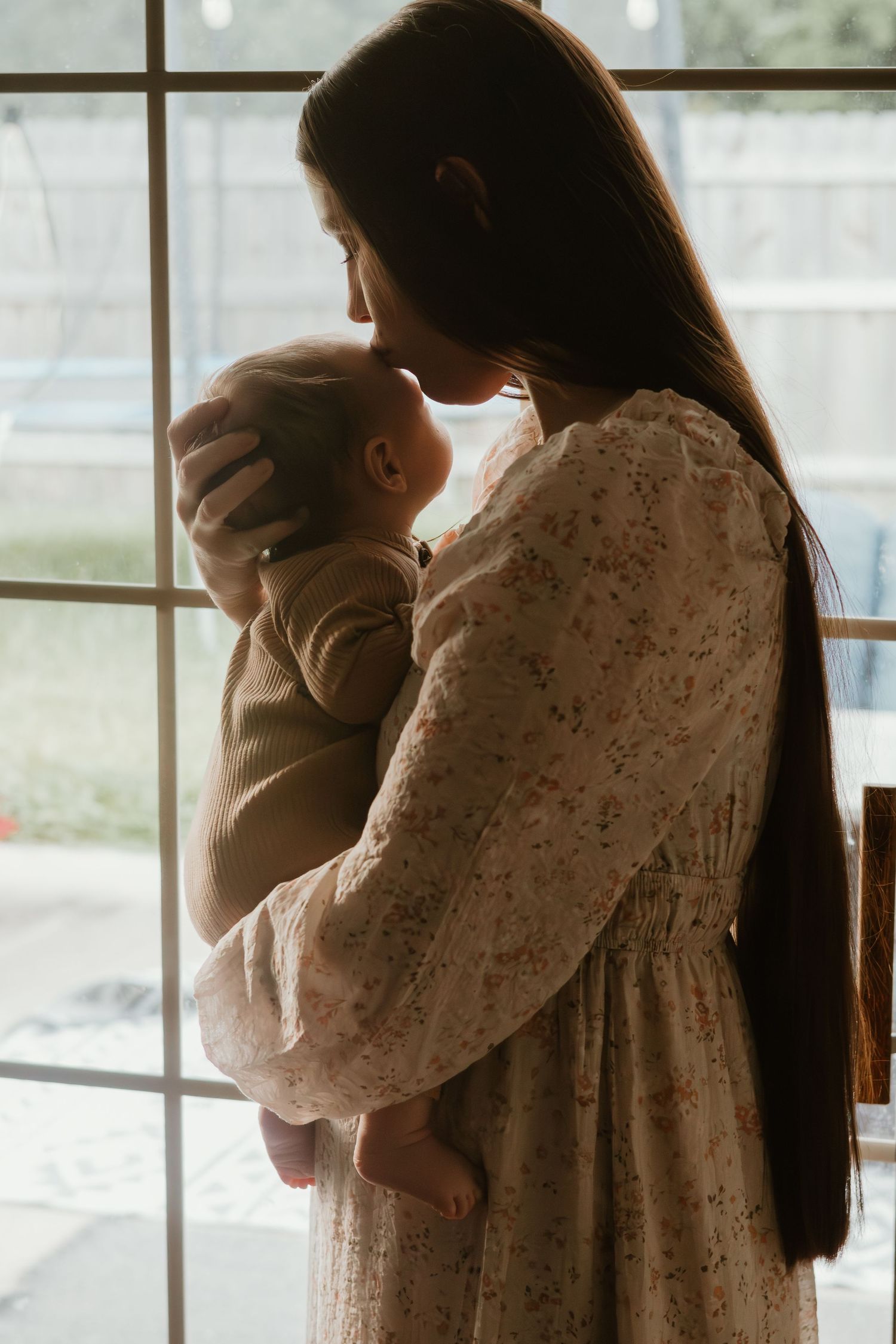 A tender moment between parent and infant silhouetted against a bright window in a floral dress.